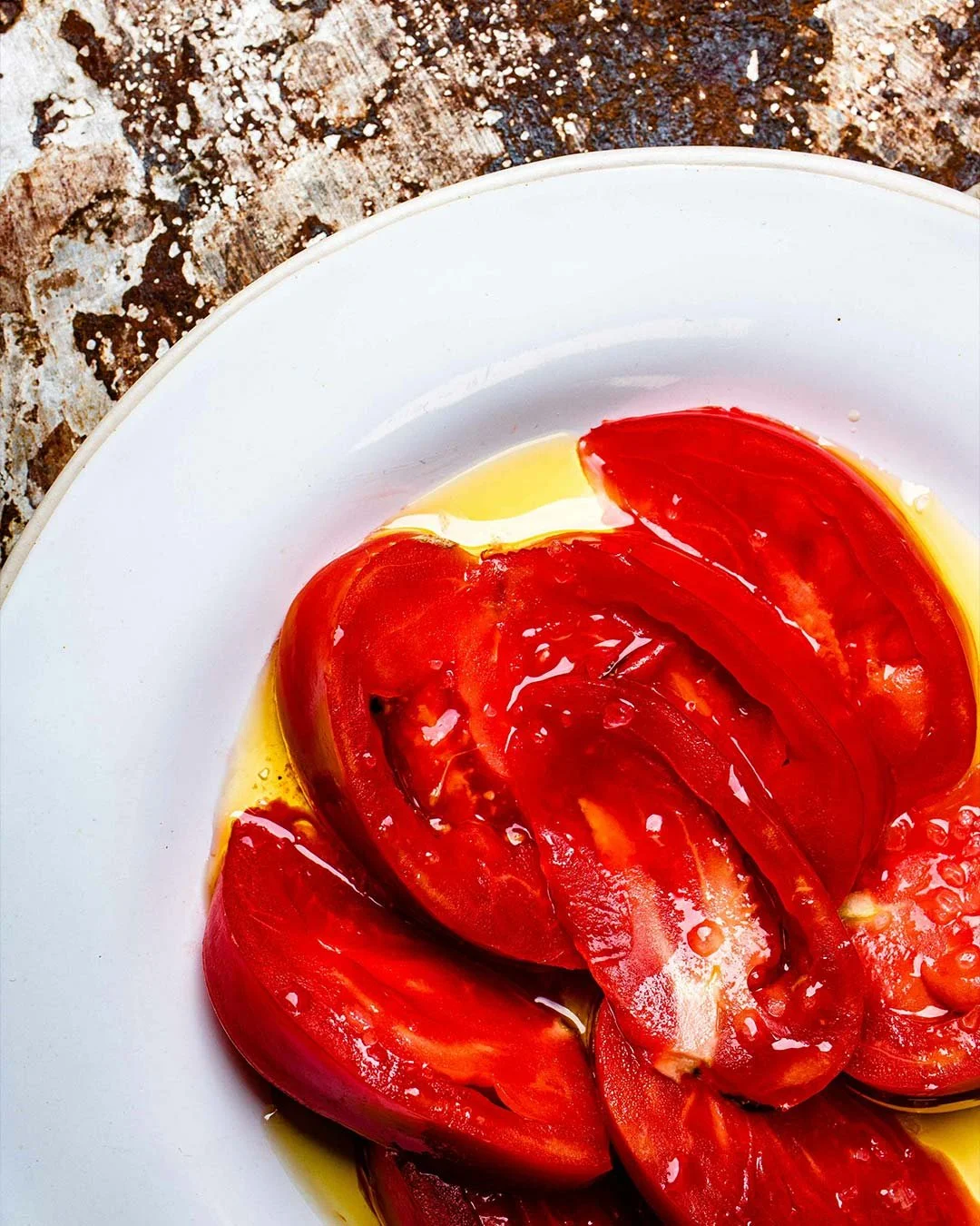 Sliced red tomatoes on a white plate with some oil or juice visible, placed on a weathered wooden surface.