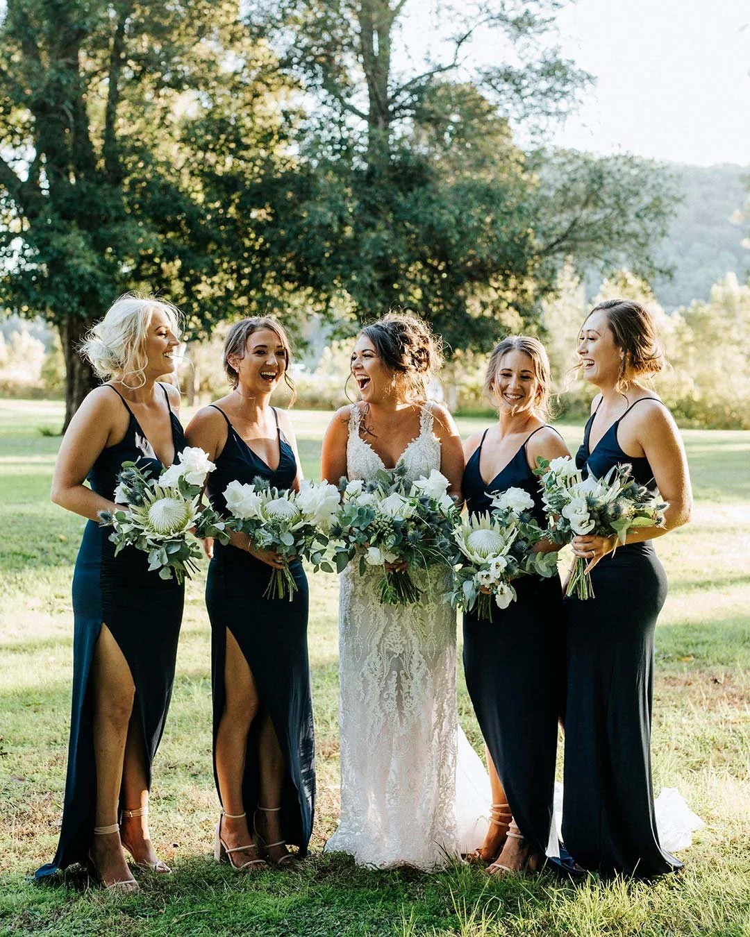A bride and her four bridesmaids standing outdoors on a grassy field with trees in the background, all smiling and holding bouquets of white flowers and green foliage.