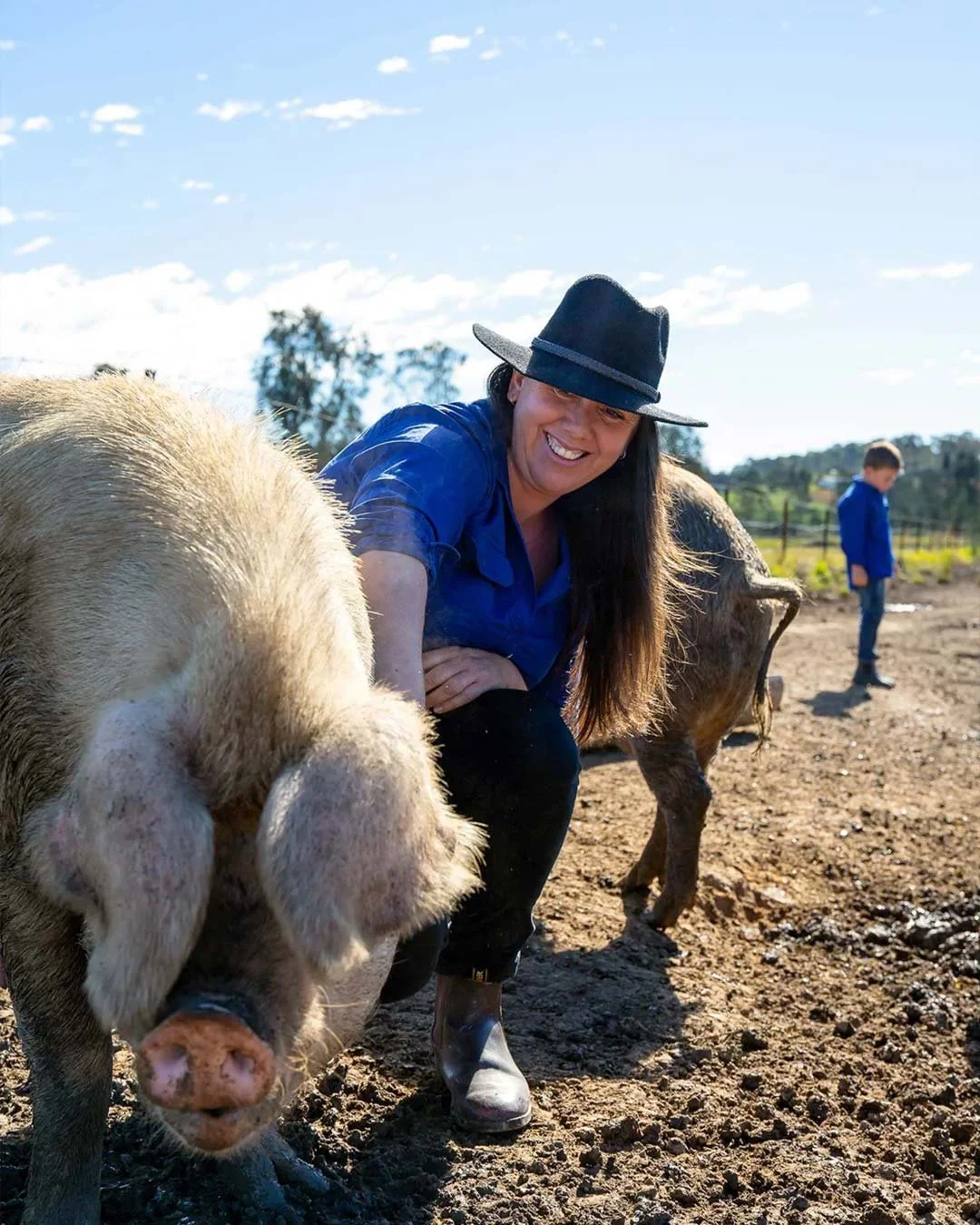 A woman in a blue shirt and black hat kneels next to a pig on a farm, smiling at the camera. In the background, a boy in a blue shirt stands near a fence and trees under a partly cloudy sky.