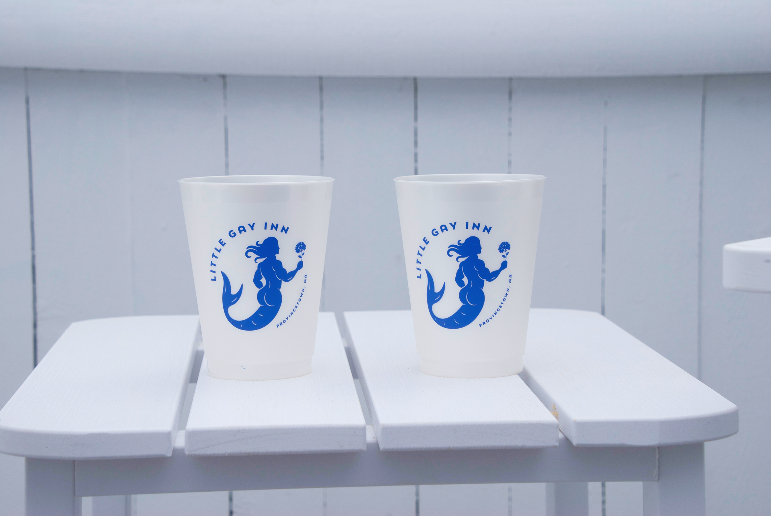 Two white cups with blue logo reading "Little Gay Inn" placed on a white surface against a light gray tiled wall.
