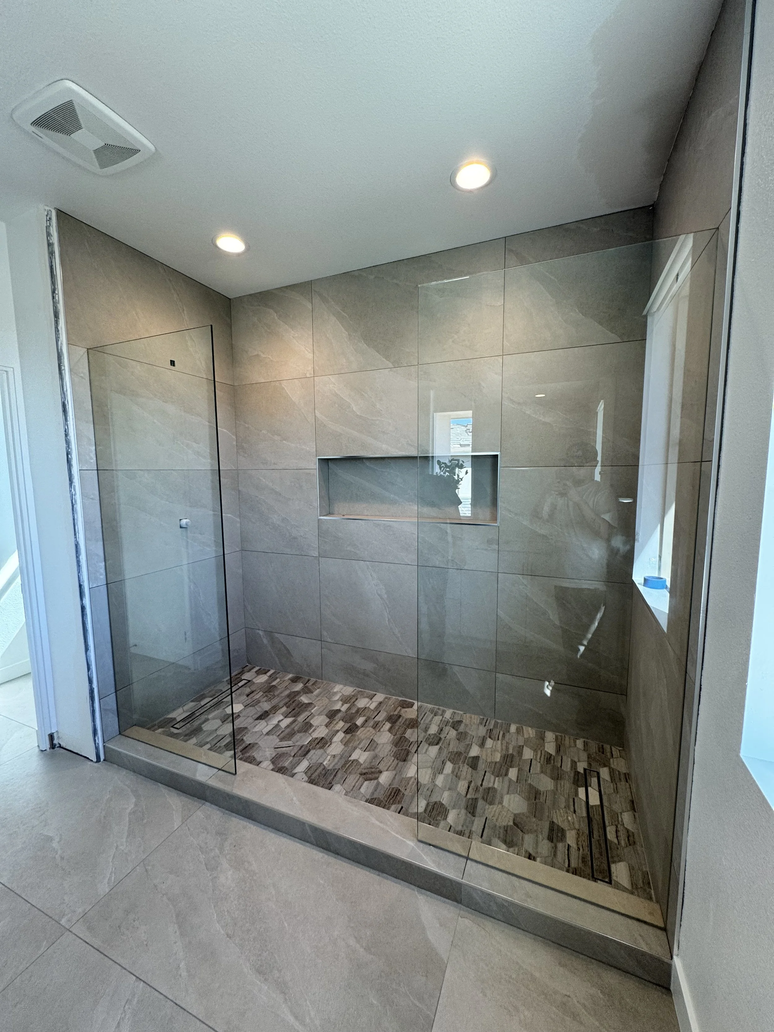 Modern walk-in shower with tan tiles, a built-in niche, and a mosaic tile floor, enclosed by clear glass panels.
