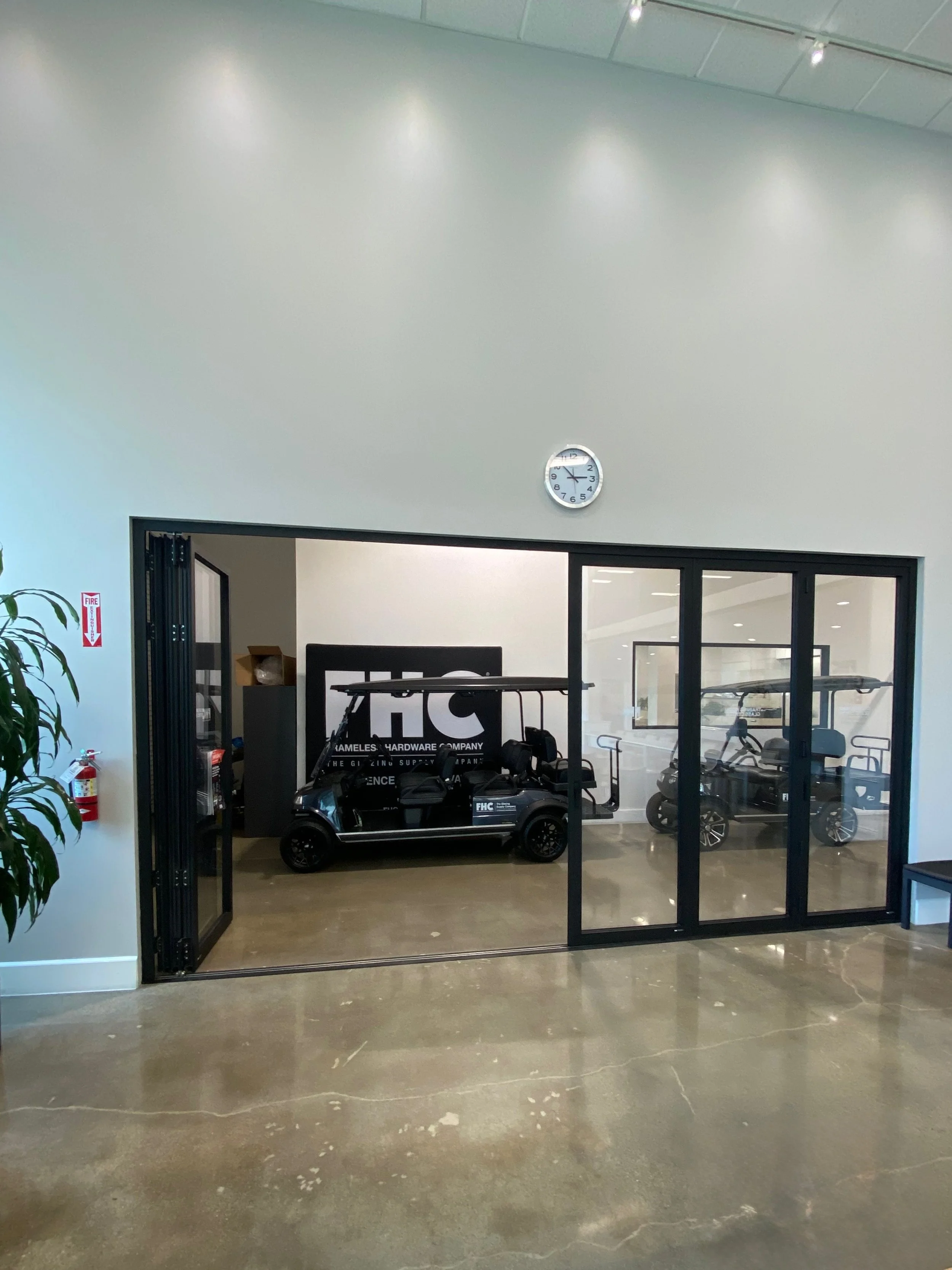 Indoor golf cart display with two carts, one in black and the other in gray, behind a glass wall in a modern building with a clock on the wall.
