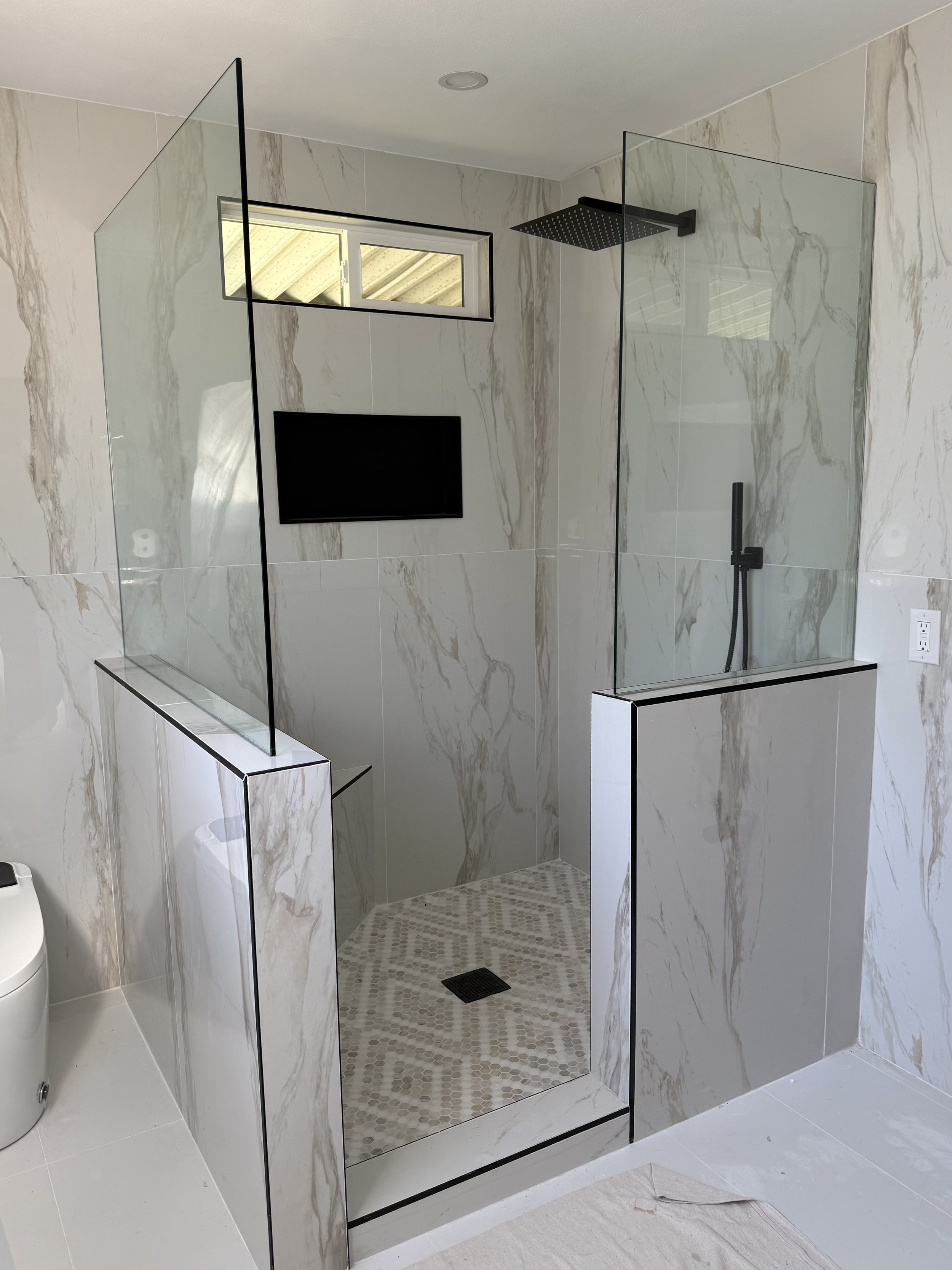 Modern bathroom with a walk-in shower enclosed by clear glass panels, black trims, a rainfall showerhead, a handheld shower, a small window, and a mounted flat-screen TV on a marble wall.