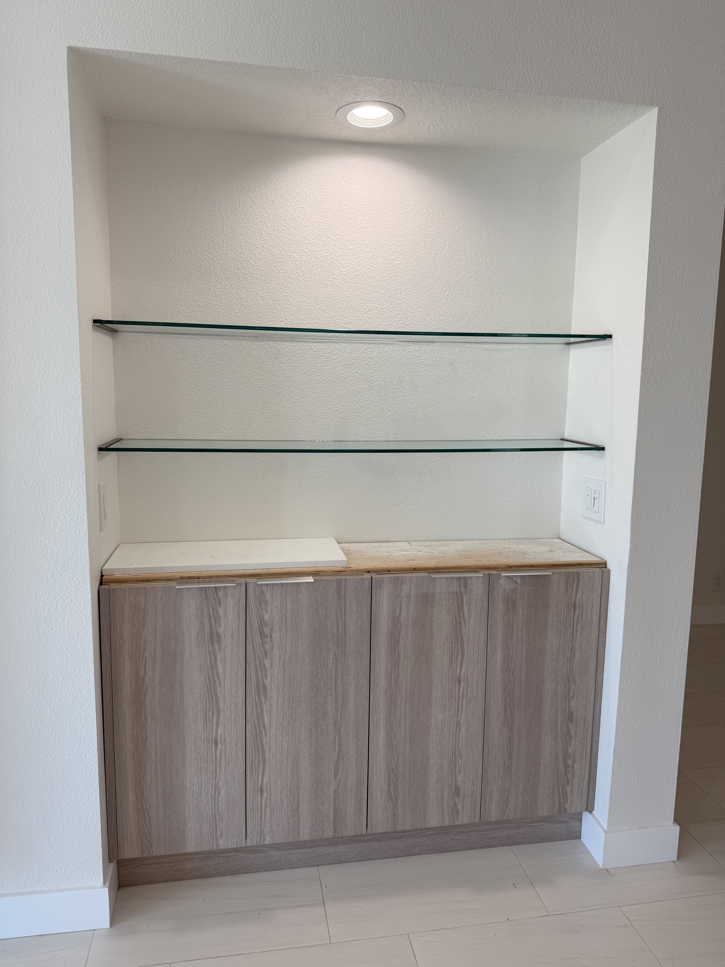 Built-in cabinet with three glass shelves above and a wooden cabinet below, in a room with white walls and light-colored flooring, illuminated by a ceiling light.