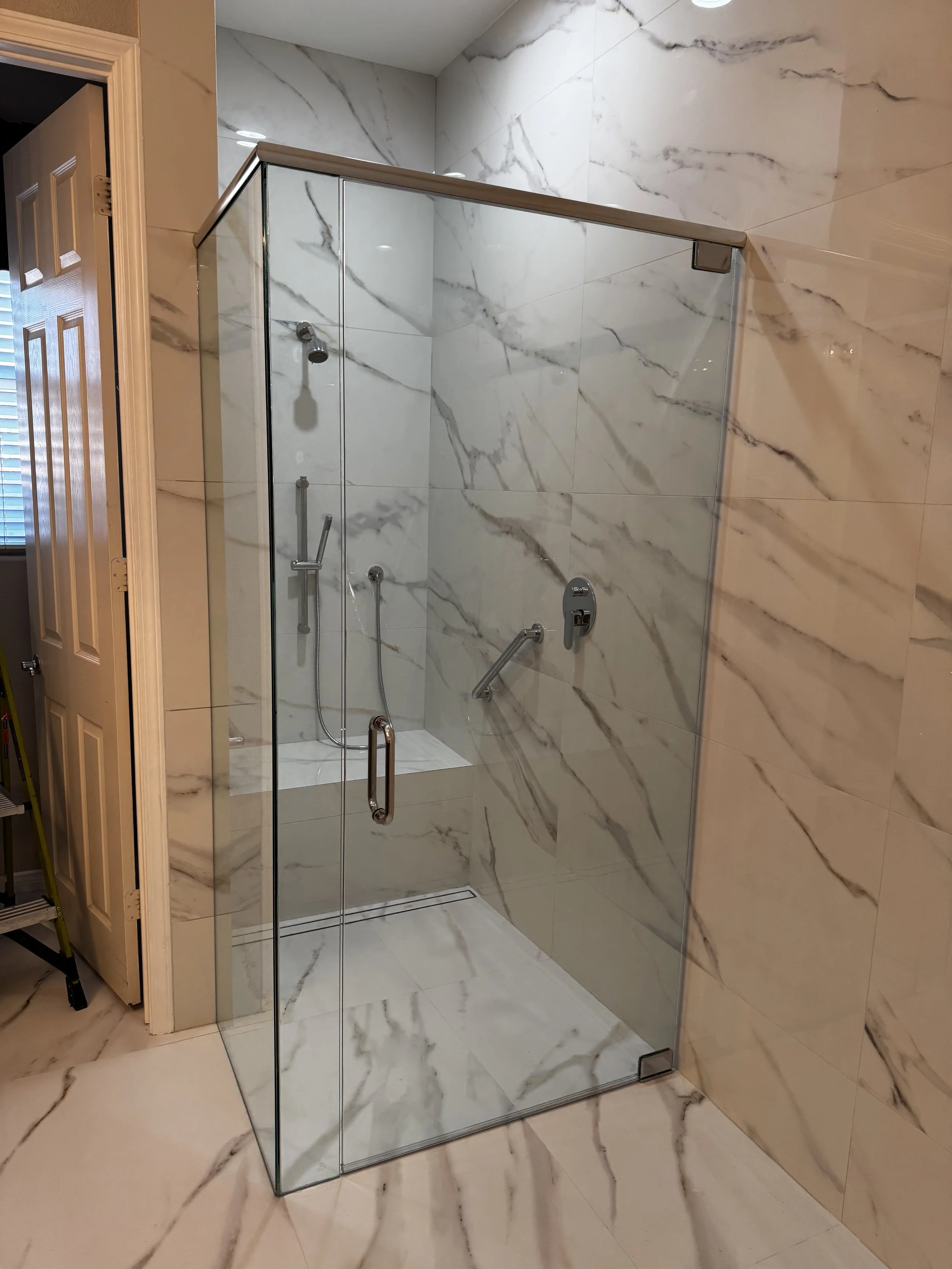 A glass shower enclosure with marble walls and flooring, featuring a stainless steel handle and fixtures, and a built-in seating area.