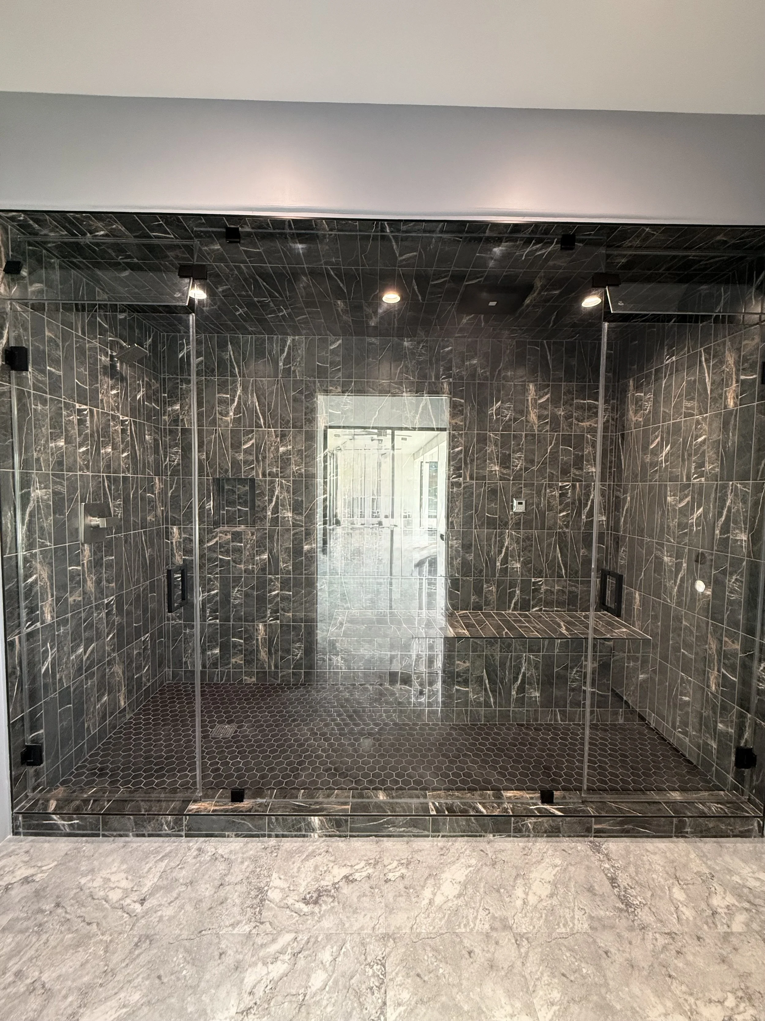 A walk-in shower with dark gray and black marble tiles on the walls and ceiling, glass doors, and a built-in bench. The floor inside the shower has small dark hexagonal tiles, and there is a window at the back of the shower.