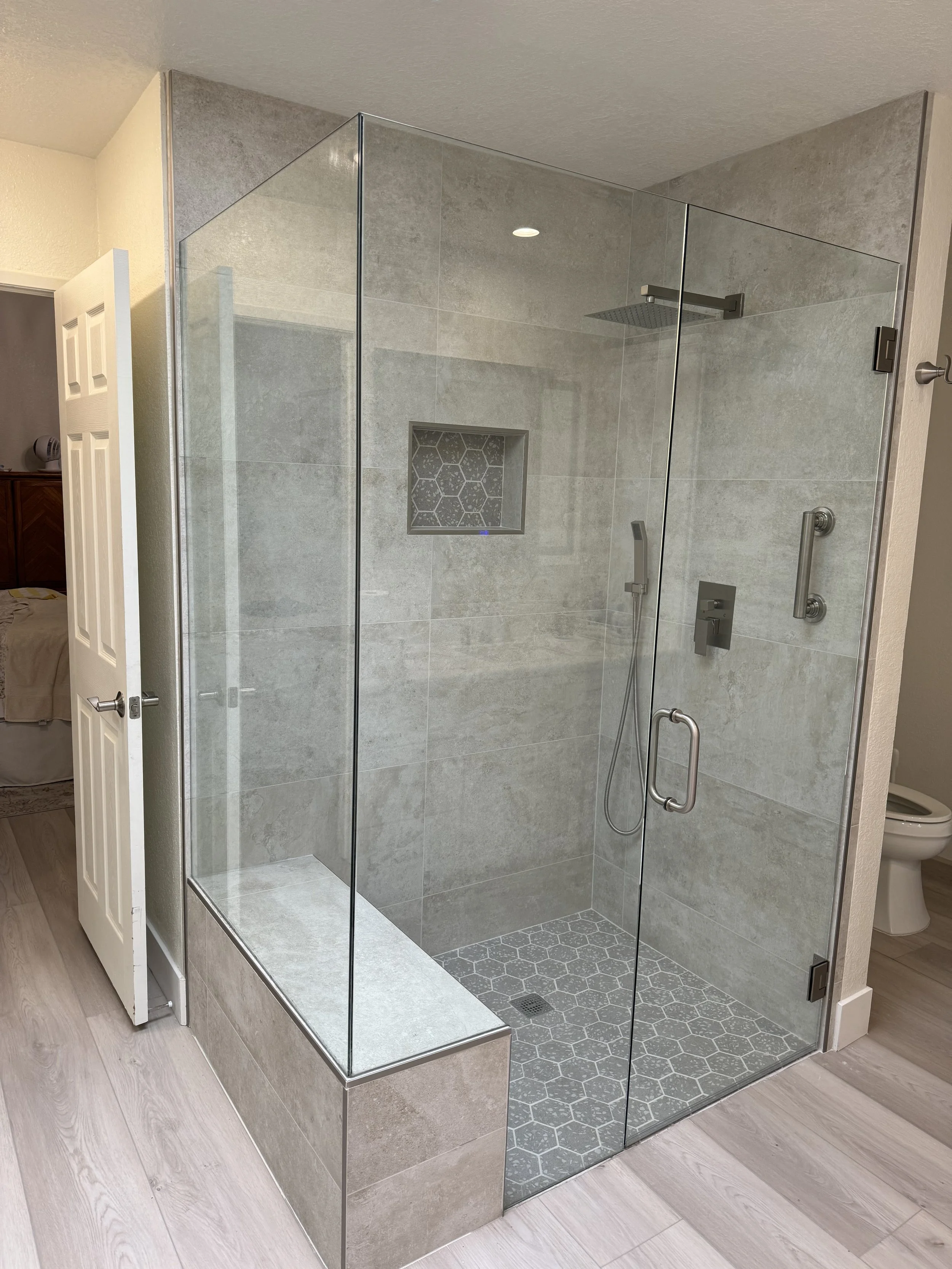 A modern walk-in shower with clear glass walls, a built-in tiled bench, gray tile walls, hexagonal gray floor tiles, a wall niche with hexagonal tile pattern, a rainfall showerhead, a handheld shower, and a grab bar. The shower is adjacent to a bathr