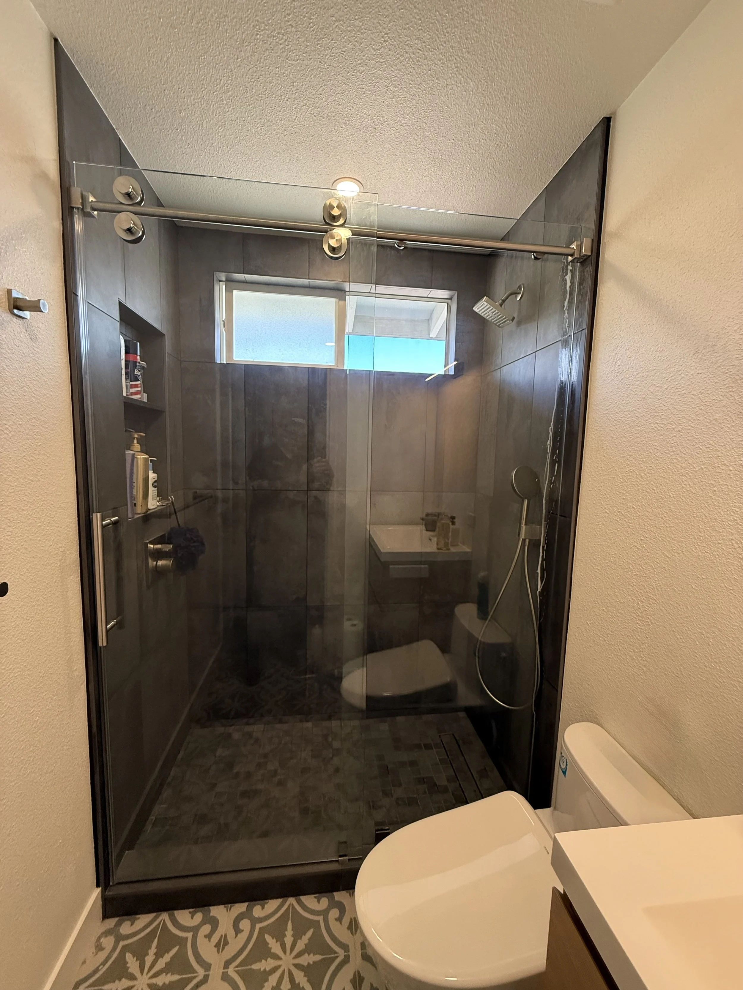 A modern bathroom with a glass shower enclosure, gray tiles, a small window, a showerhead, a handheld sprayer, a toilet, and a patterned tile floor.