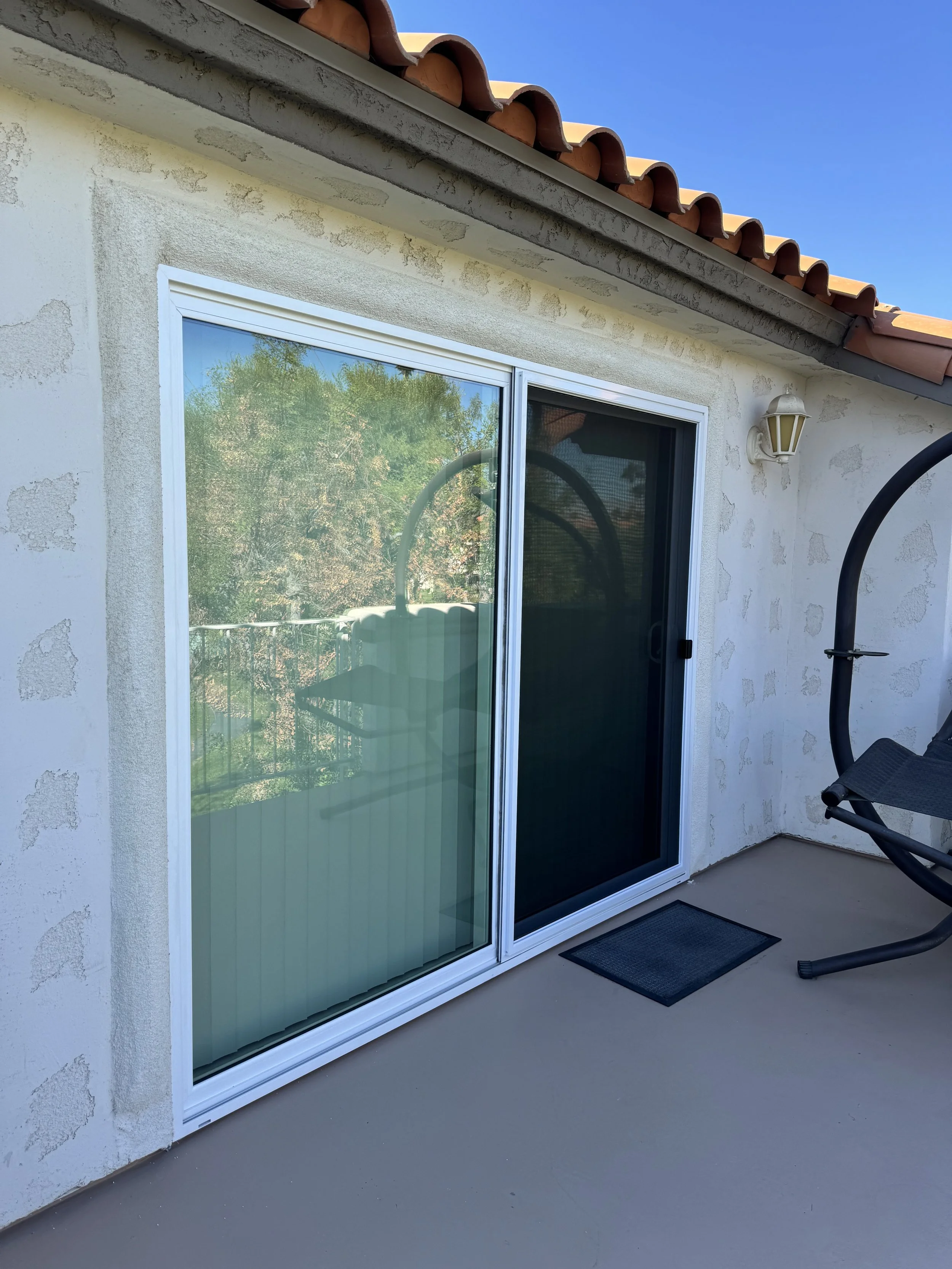 Exterior view of a balcony with a sliding glass door, a small black doormat, a patio chair, and a wall-mounted lantern light fixture. The balcony has a beige concrete floor and a tiled roof with curved terracotta tiles. The wall outside appears to be