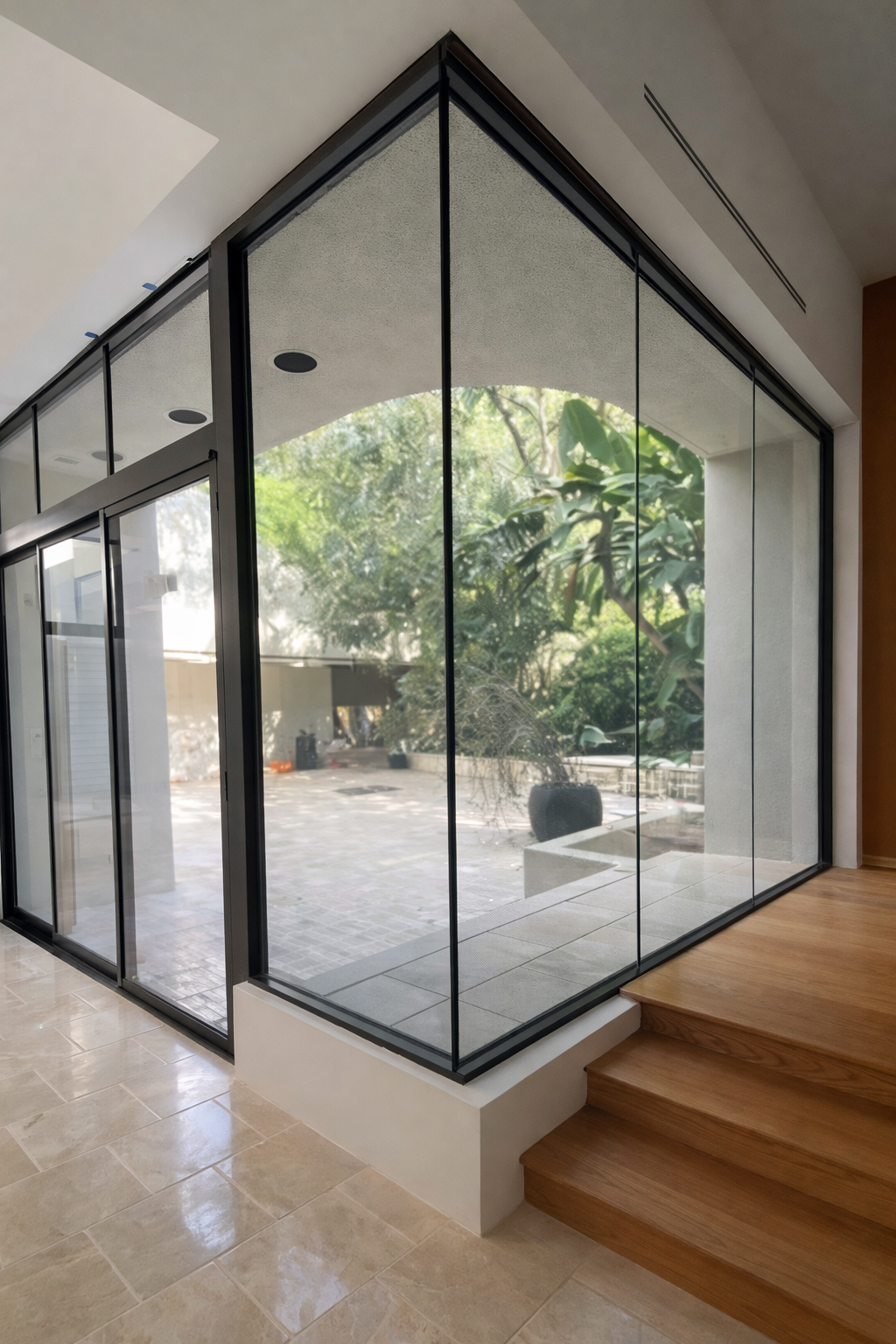 The image shows a corner of a modern interior space with large floor-to-ceiling glass windows and sliding glass doors framed in black metal, revealing a lush outdoor area with trees and plants. There are light beige tiled floors and wooden stairs lea