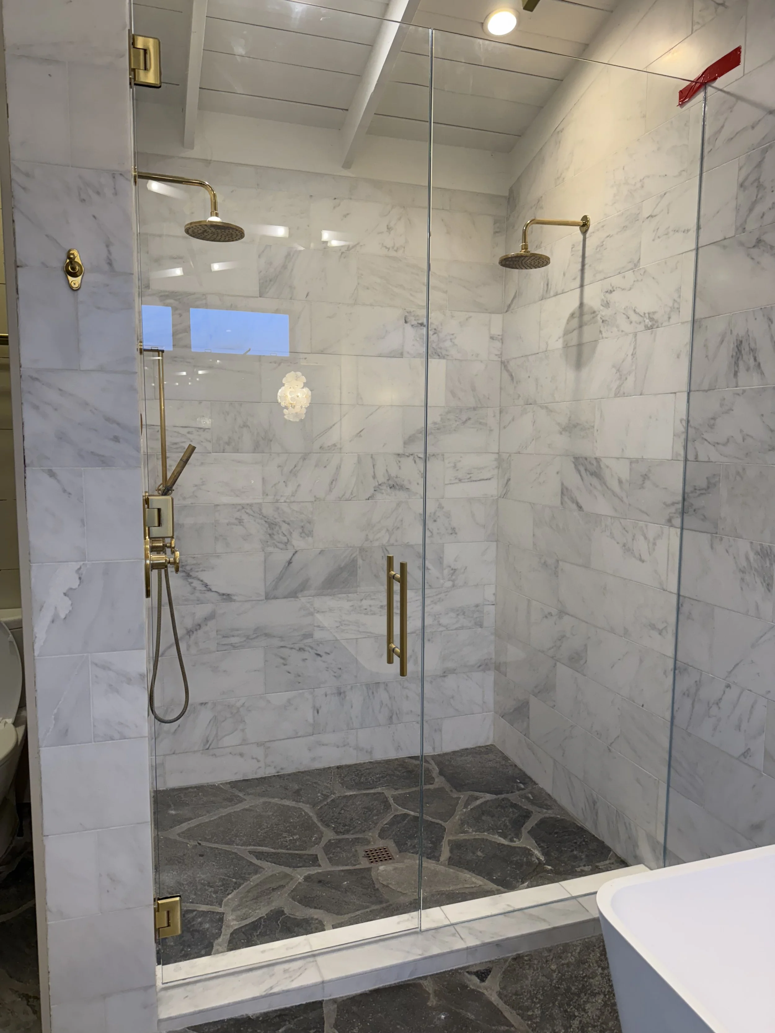 A modern walk-in shower with glass doors, white marble walls, two overhead rainfall shower heads, and a dark stone tiled floor with a drain in the center.