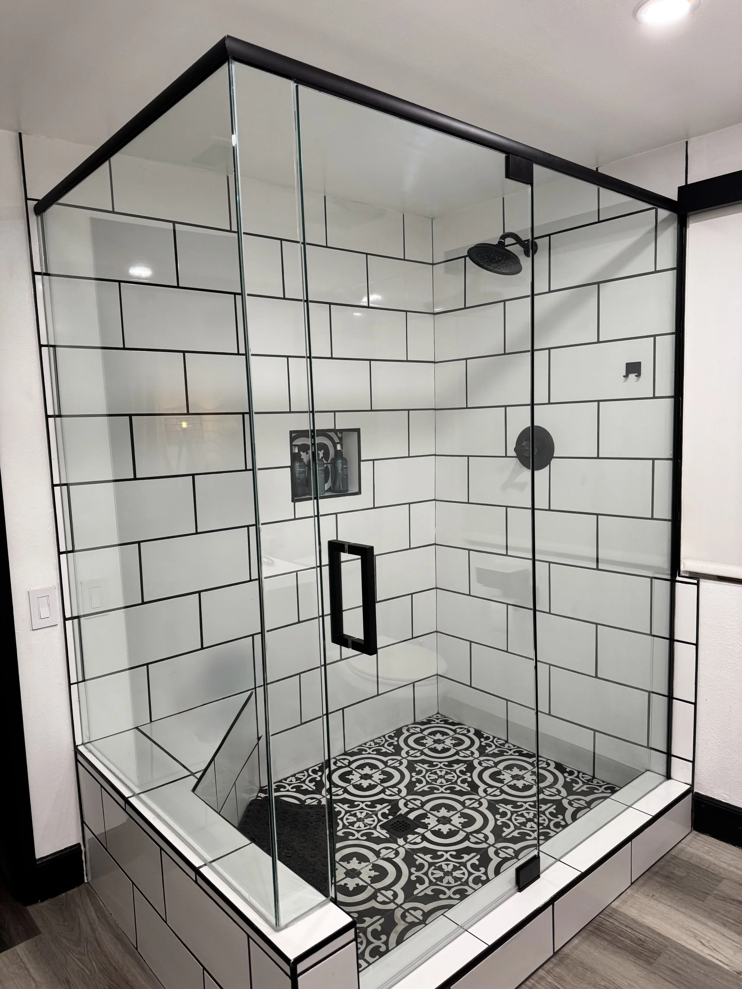 A modern glass shower enclosure with black fixtures, patterned floor tiles and white subway tiles on the walls with black grout.