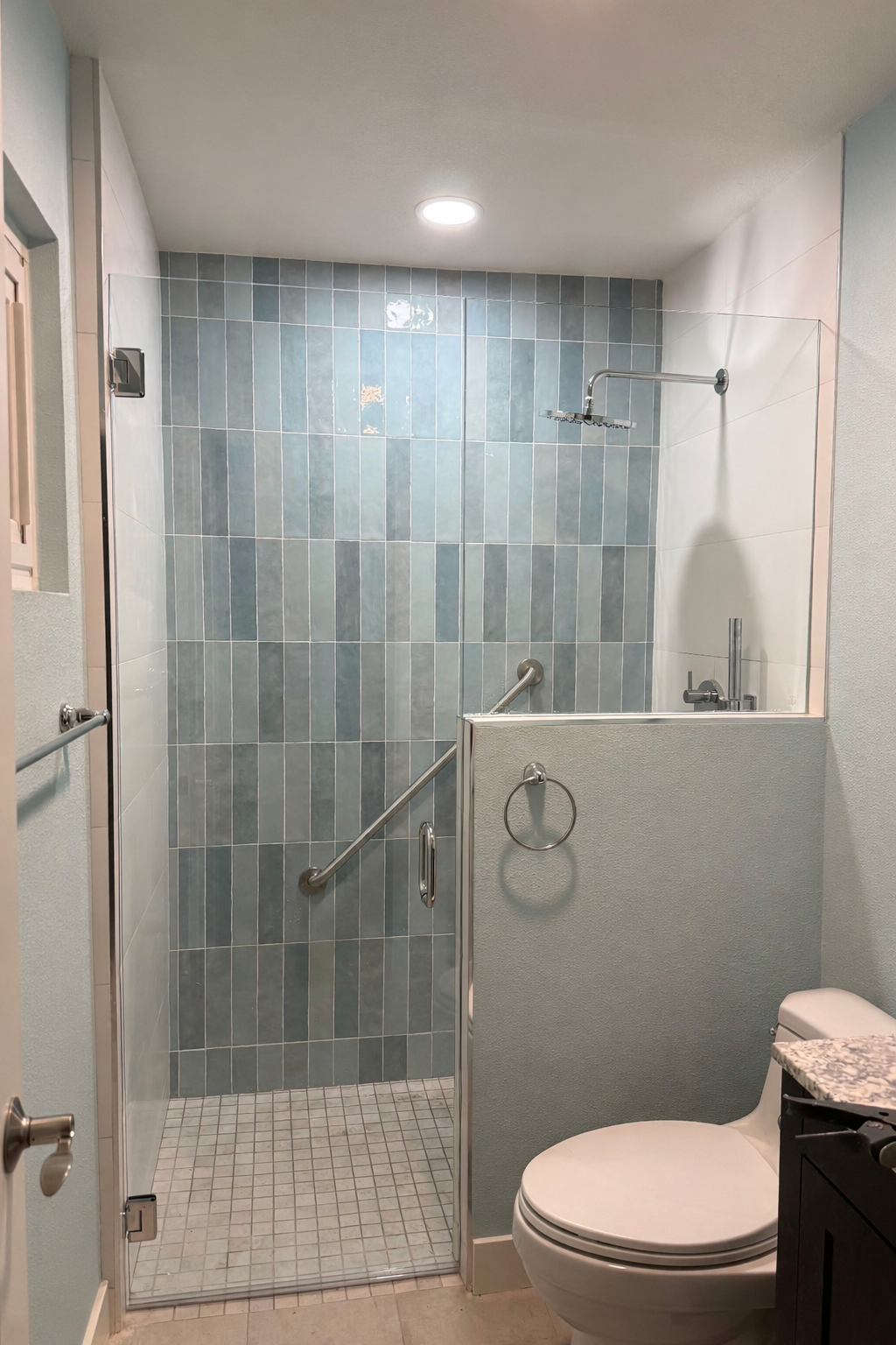 A walk-in shower with blue tiled walls, a glass door, and a showerhead. Adjacent to the shower is a toilet with a granite countertop.
