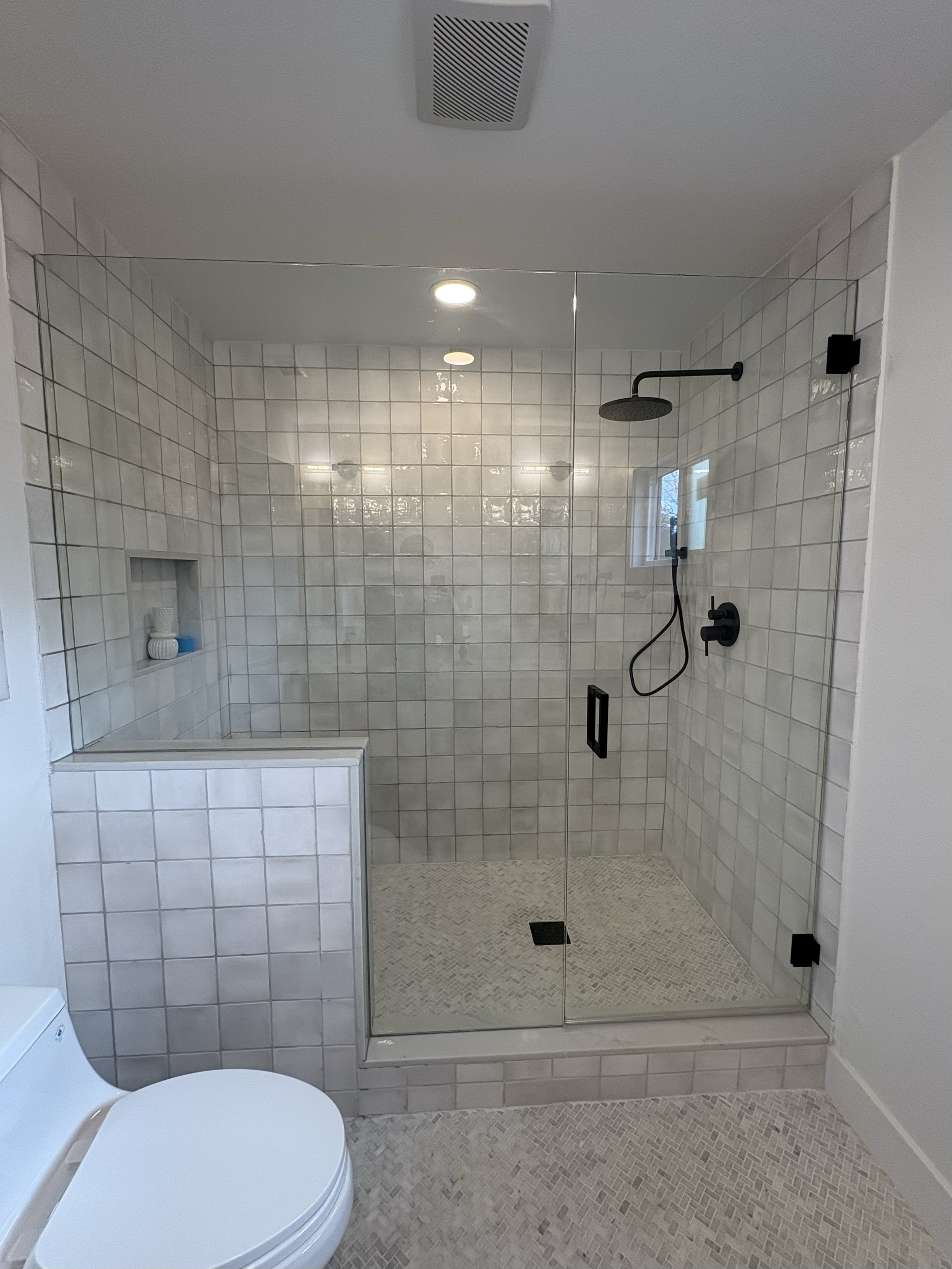 Modern walk-in shower with glass doors, black fixtures, white tiled walls, small mosaic tile floor, and a built-in niche for toiletries.