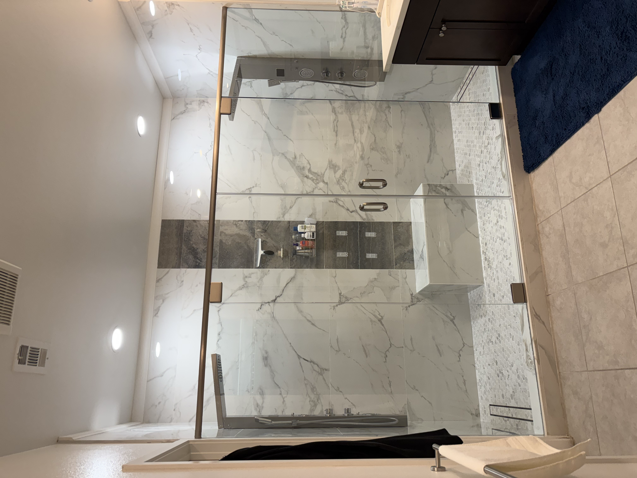Modern bathroom with a glass-walled shower, marble tiles, a small built-in shower seat, and a dark blue rug on beige tile floor.