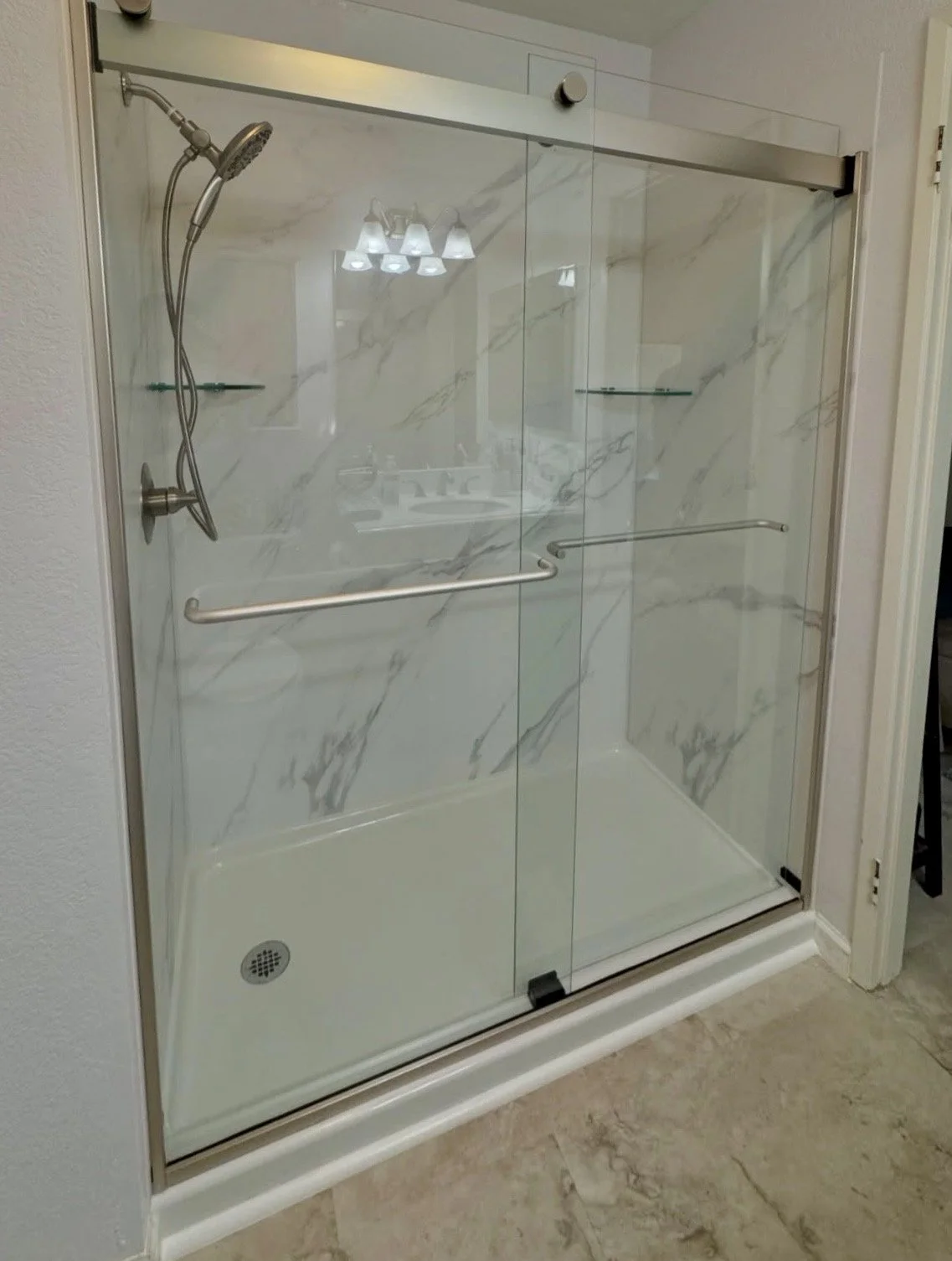 A walk-in shower with glass sliding doors, a handheld showerhead, marble-patterned walls, a glass shelf, and a black drain; beige tile floor outside.