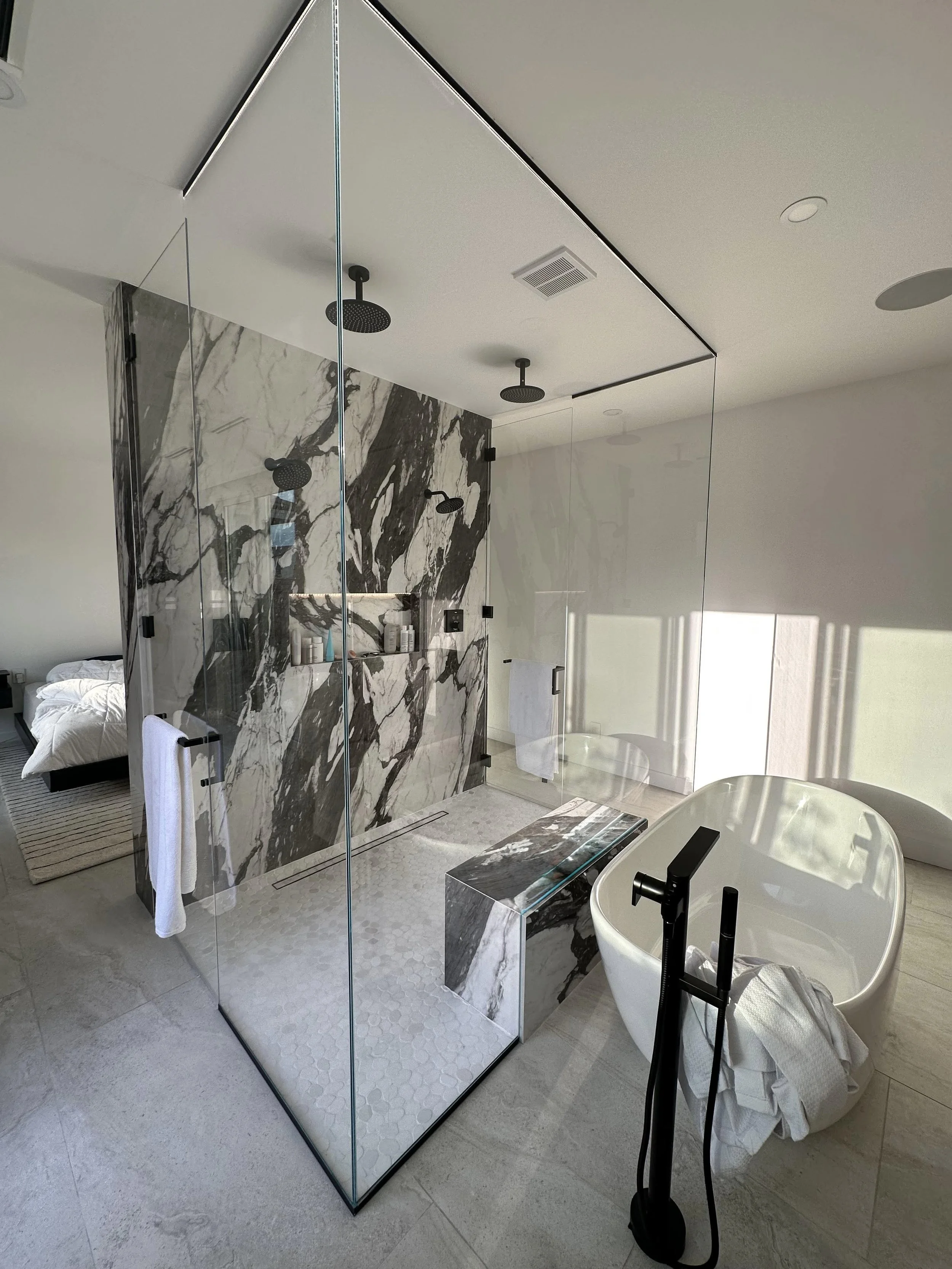 Modern bathroom with a glass-enclosed shower area featuring black showerheads and shelves with toiletries. There is a black and white marbled wall inside the shower. Outside the shower, there is a white freestanding bathtub with a black faucet, near 
