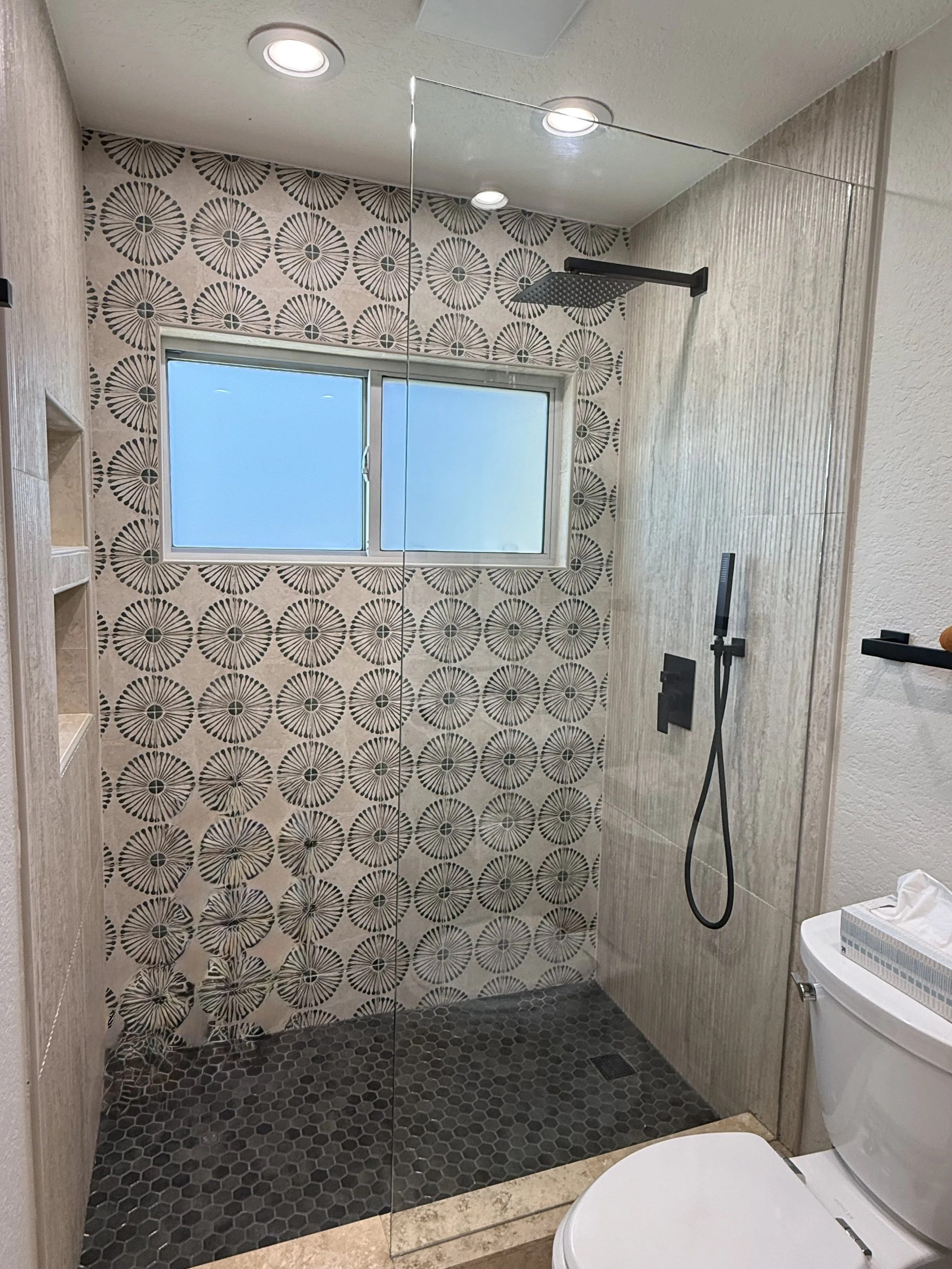 Modern bathroom with a walk-in shower featuring geometric patterned tiles on the wall, a black overhead showerhead, a handheld shower on a black bar, a small window, and a white toilet.