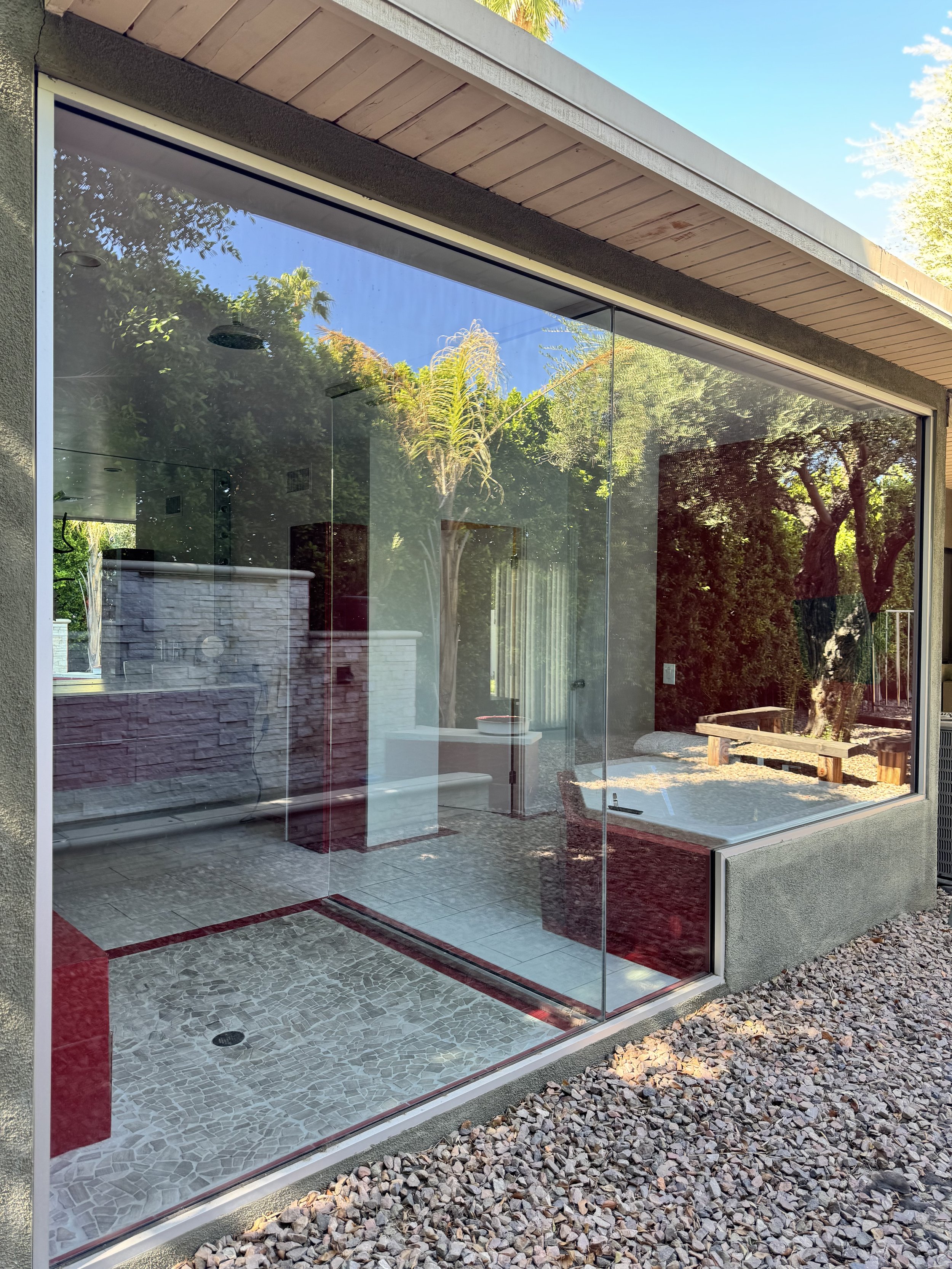 A modern built house with a large glass window, providing a view into a room with a gray and red interior, and an outdoor area with benches and trees.