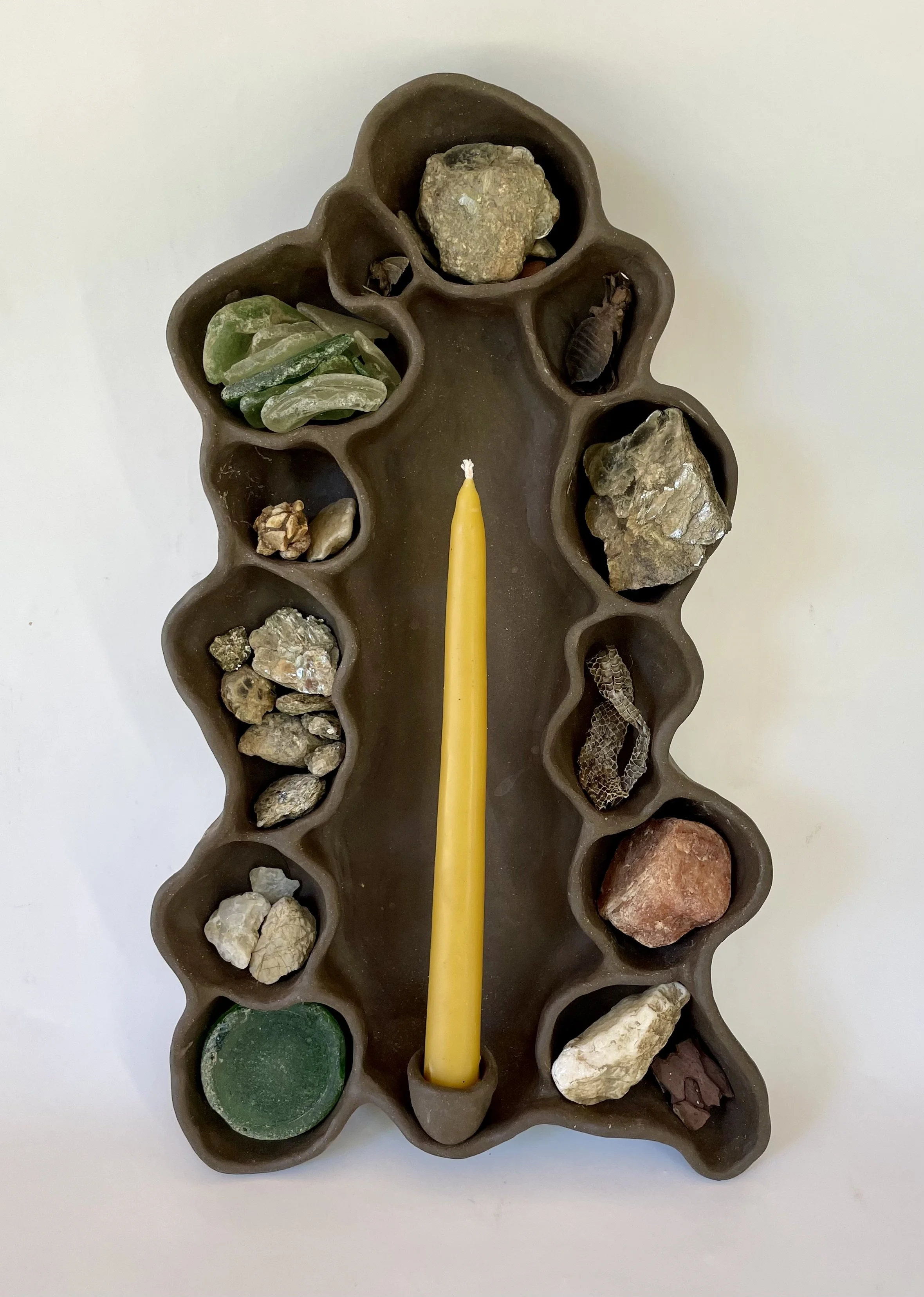 A brown candle holder with small compartments filled with various rocks and minerals, and a yellow candle in the center.