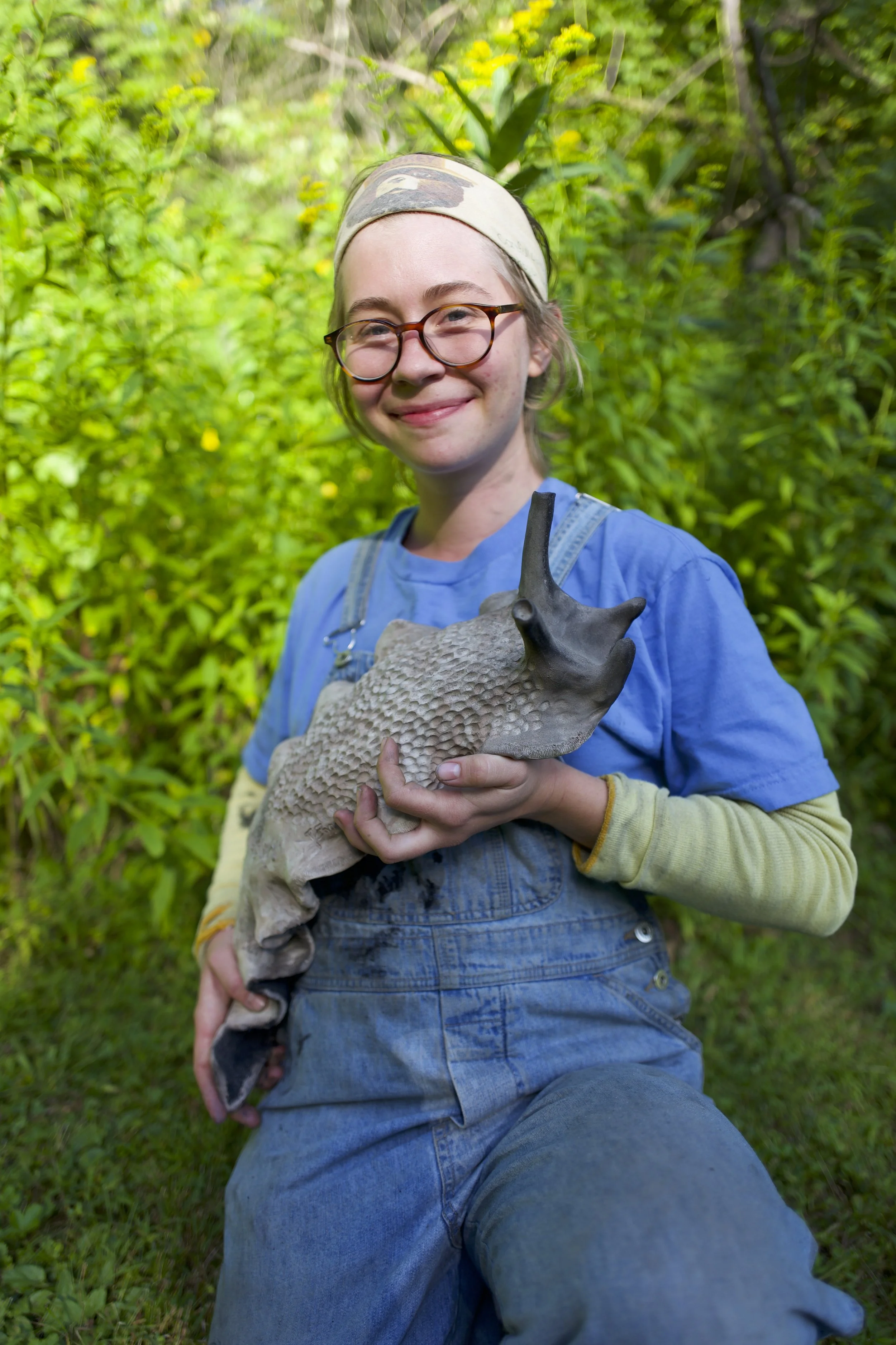 A young person with glasses and a headscarf kneeling in greenery, smiling, holding a ceramic slug.