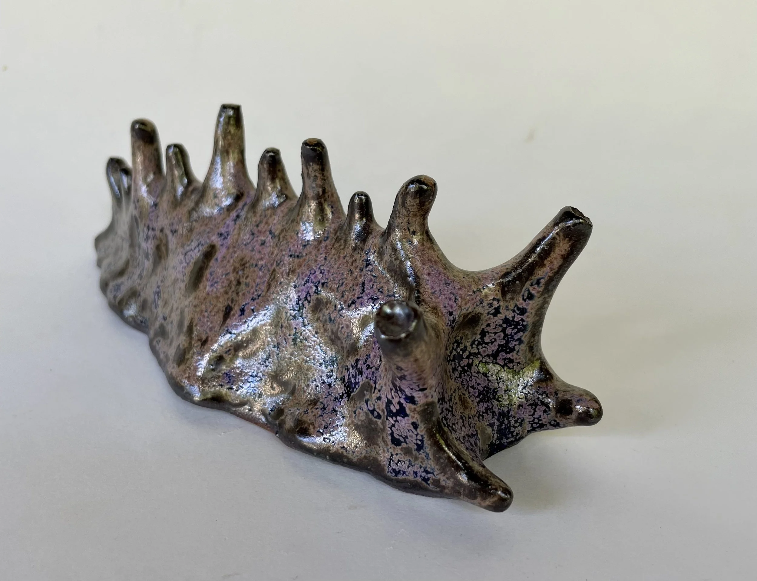 A ceramic sculpture resembling a sea slug with a series of dorsal spines or horns along its back, painted with a metallic finish and decorative patterns in pink, purple, and black.