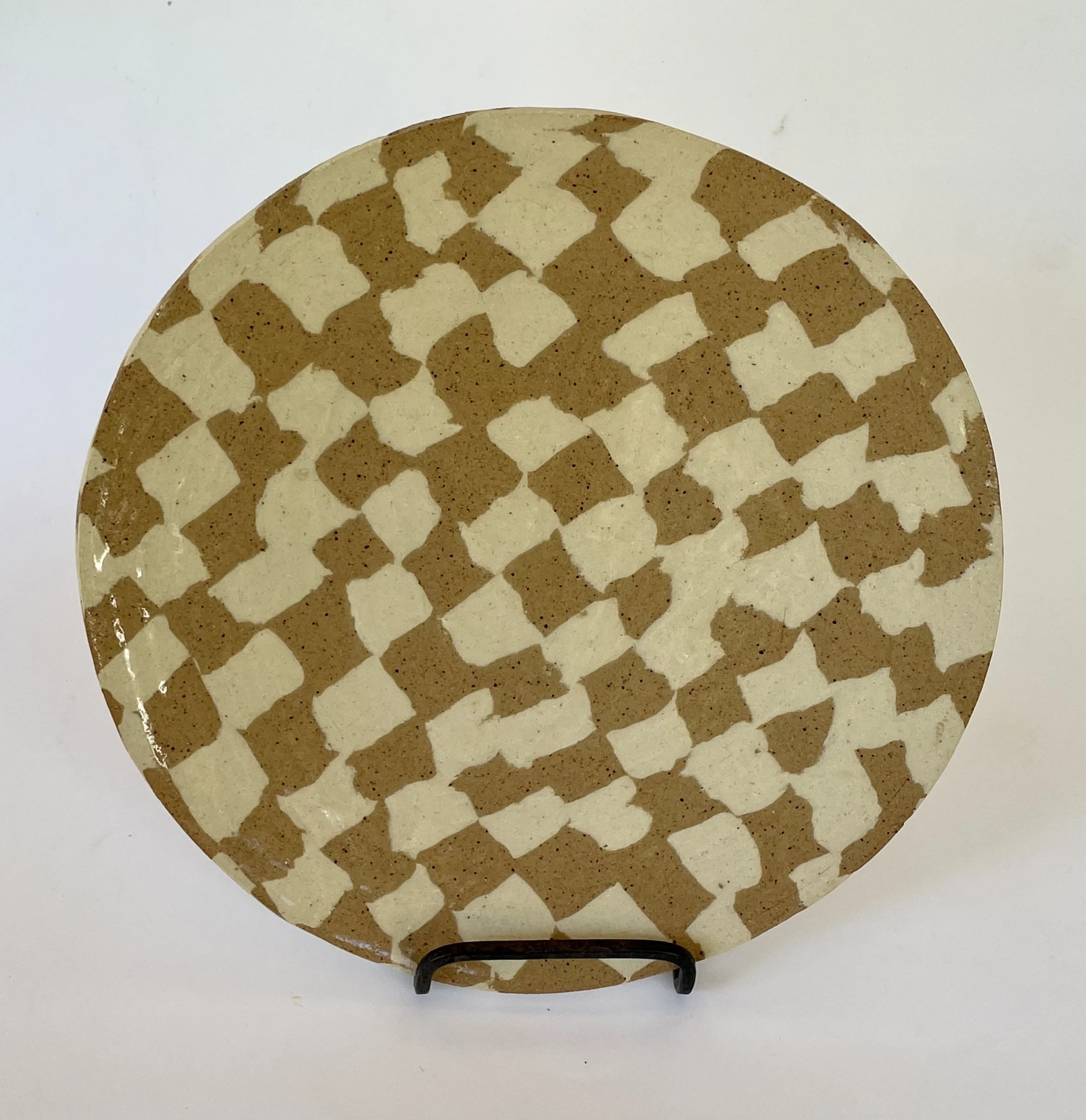 Decorative round ceramic plate with a beige and brown checkerboard pattern, displayed on a black stand against a white background.