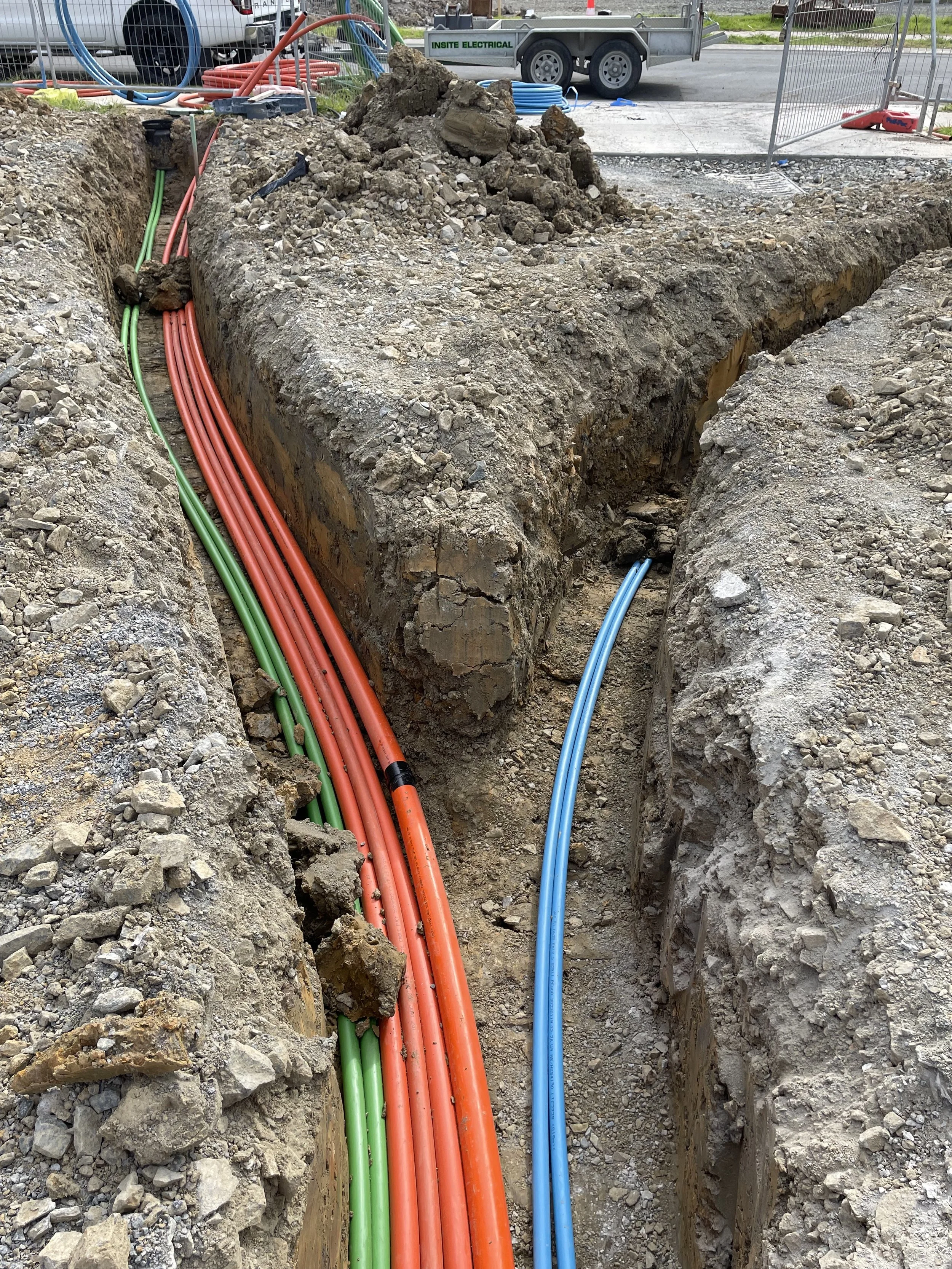 Construction site with utility pipes: green and red pipes laid in a trench, with blue pipe only partially visible, surrounded by dirt and rocks, and construction equipment and vehicles in the background.