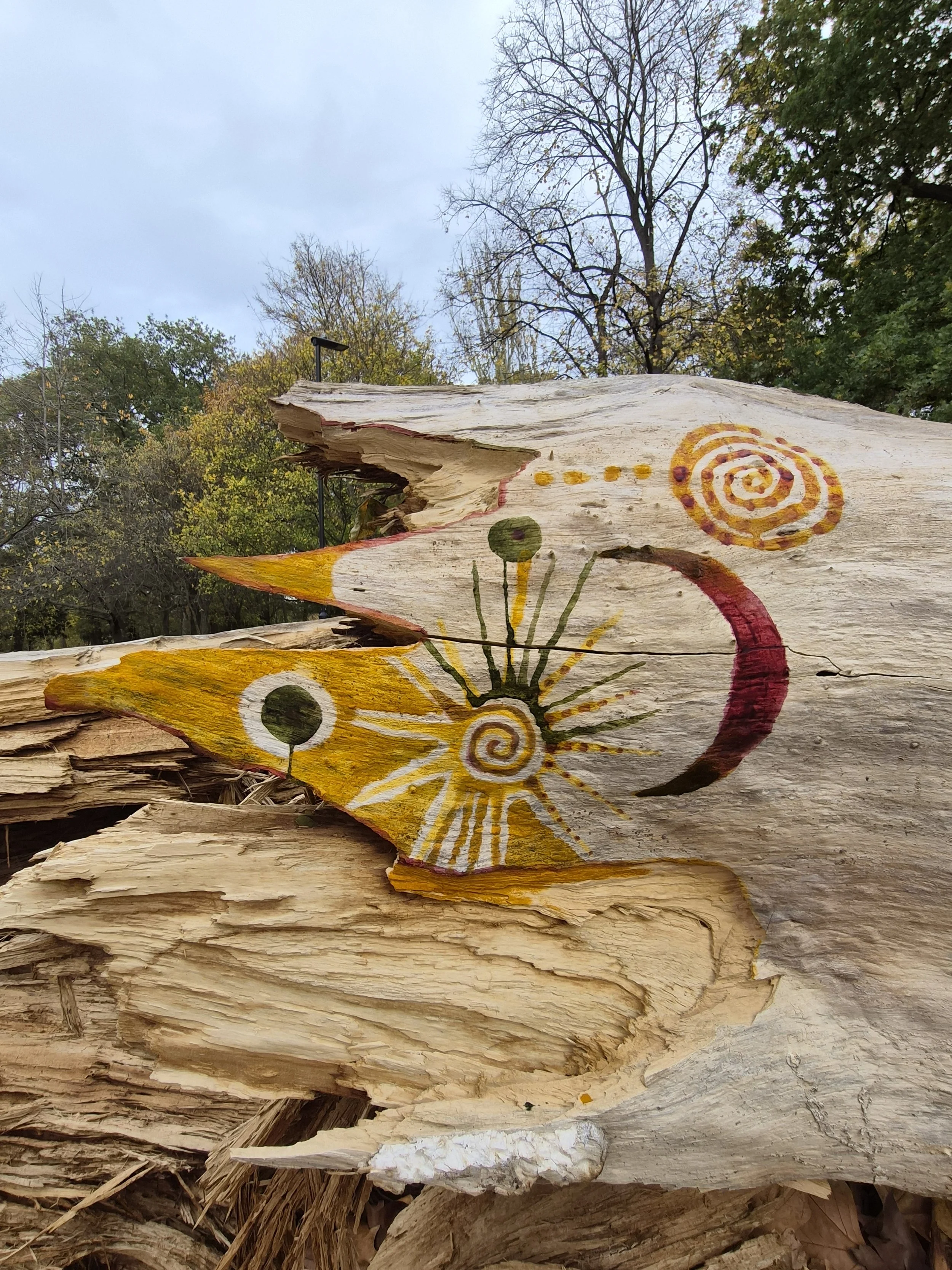 Painted abstract design on a weathered piece of wood with trees in the background.