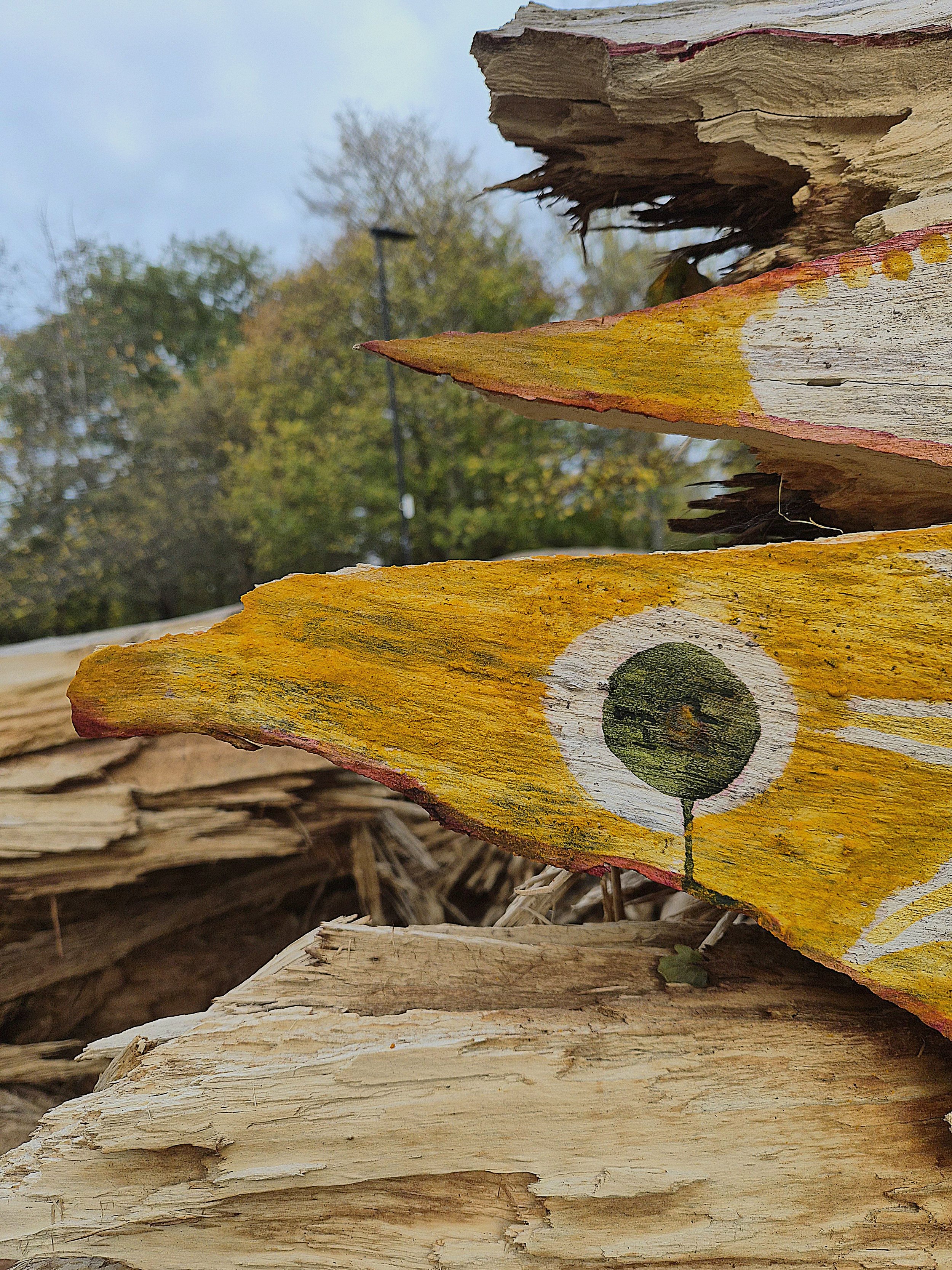 Close-up of stacked, weathered wood with yellow and red paint, featuring a painted eye design, with trees and sky in the background.