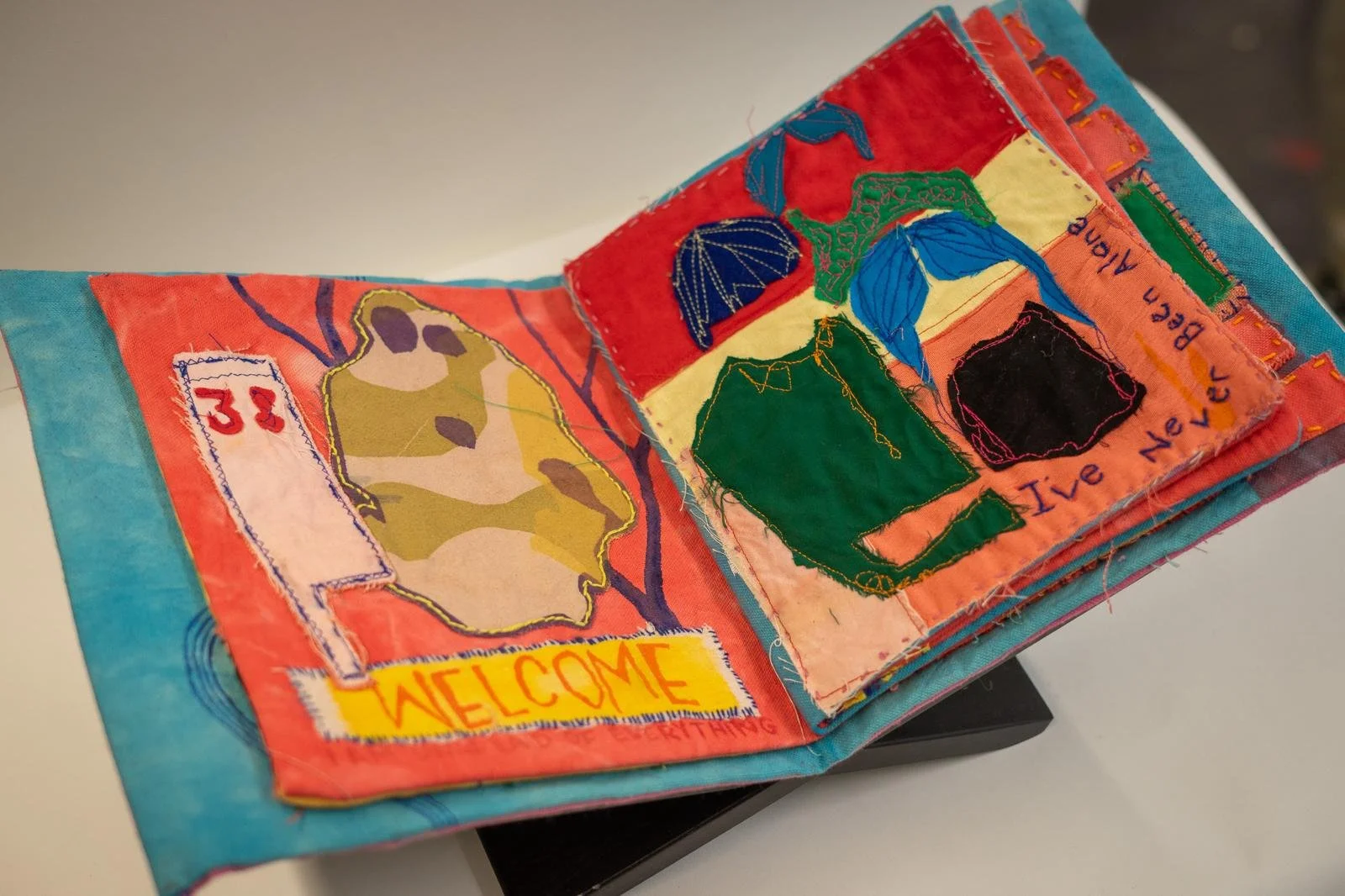 Open fabric art journal with colorful embroidered patches, drawings, and text, including a yellow 'WELCOME' patch and patches with abstract shapes and words like 'BEACH' and 'LIVE NEVER AGAIN'.