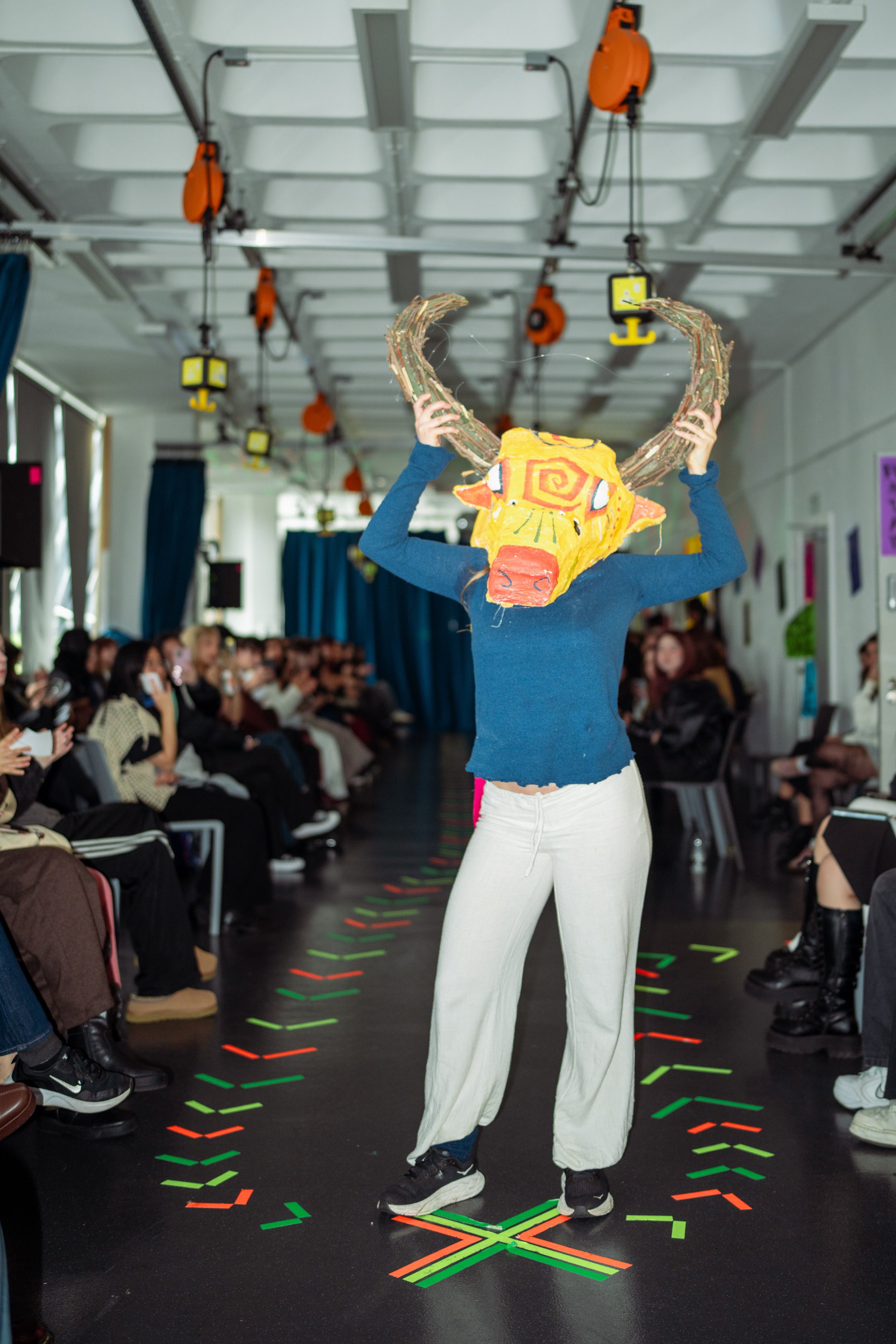 Person on a runway wearing a lion mask with large antlers, surrounded by an audience.
