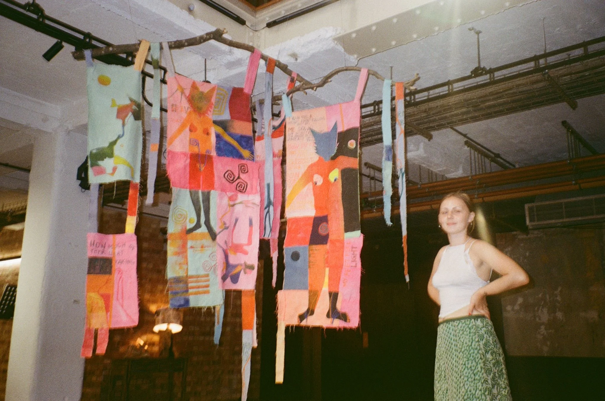 A young woman standing next to colorful fabric art hanging from a branch in an arts space or gallery.