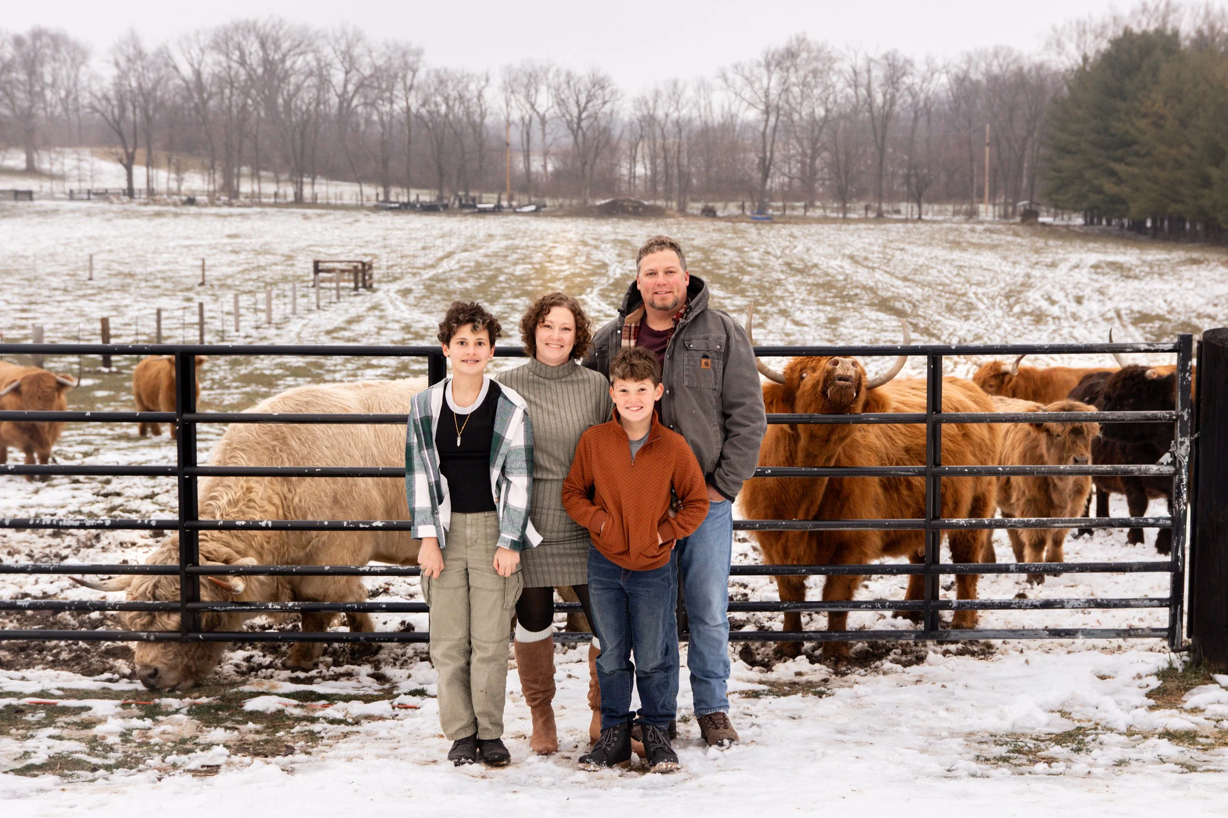 Family of four posing in front of a farm fence with bison and snow-covered field behind them