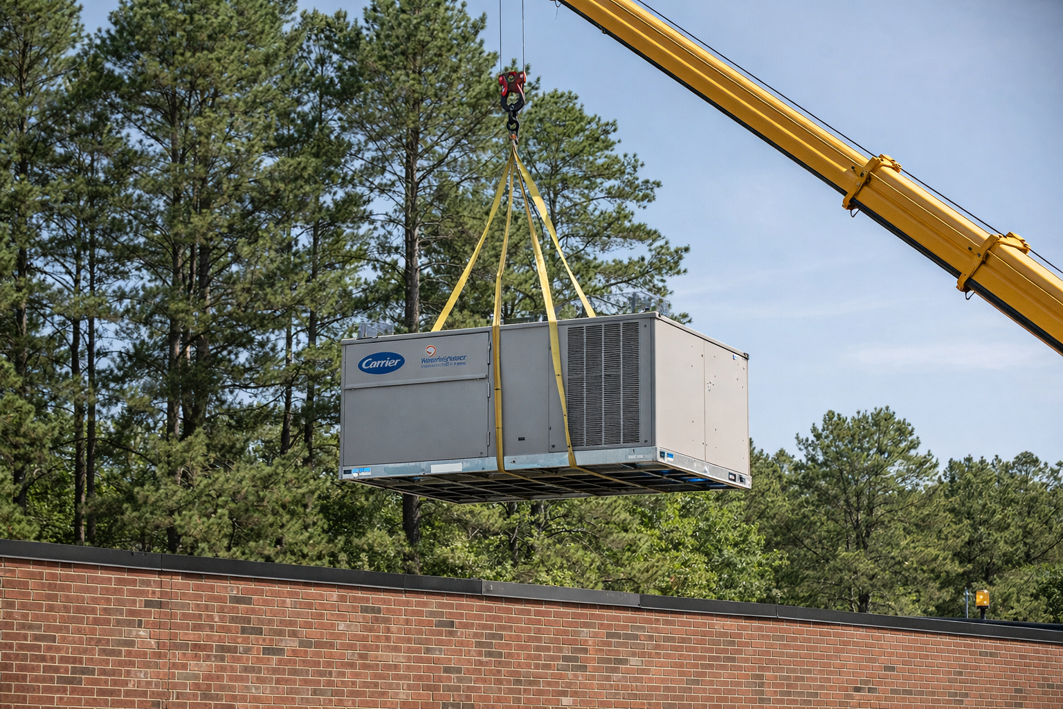 A construction crane lifting a large air conditioning unit above a brick building with green trees in the background.