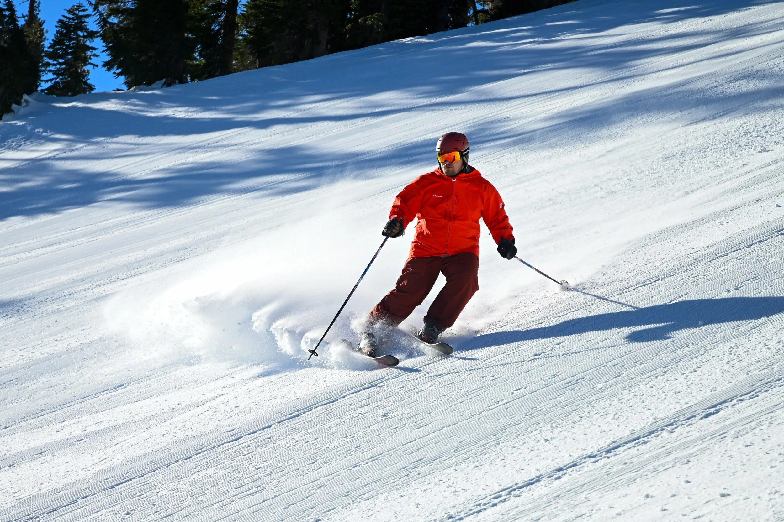 A skier in red jacket and brown pants skiing down a snow-covered slope with trees in the background.