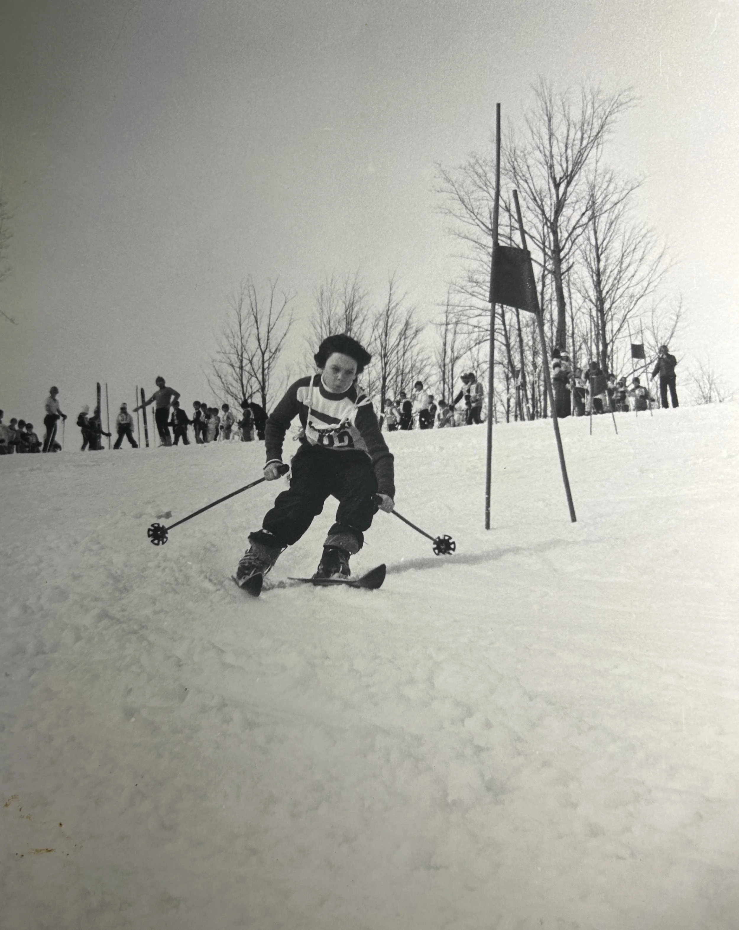 A young Dr. Russell Petrie in winter clothing skiing on a snowy slope at a ski race with spectators and other skiers in the background, leafless trees, and ski poles on the snow.