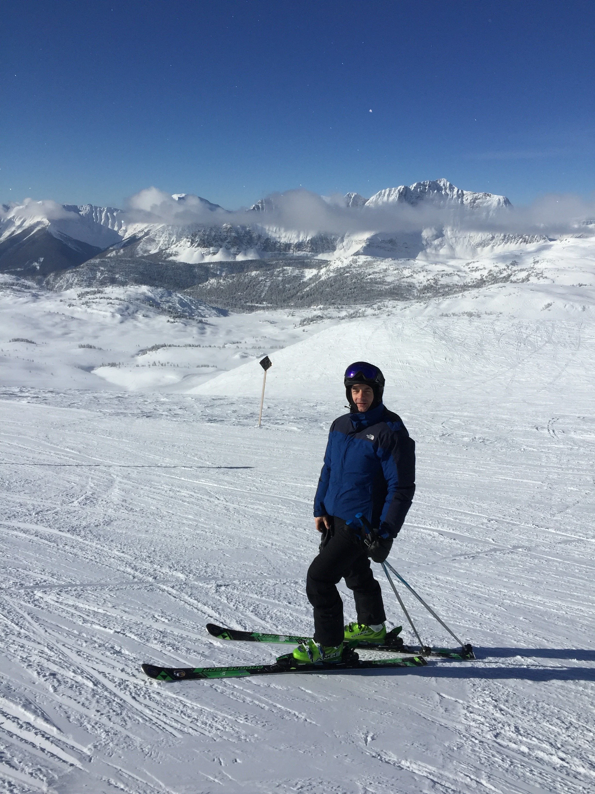 Dr. Russell Petrie wearing a blue and black ski jacket, black pants, and skiing gear, standing on a snowy mountain slope with a backdrop of snow-covered mountains and a clear blue sky.