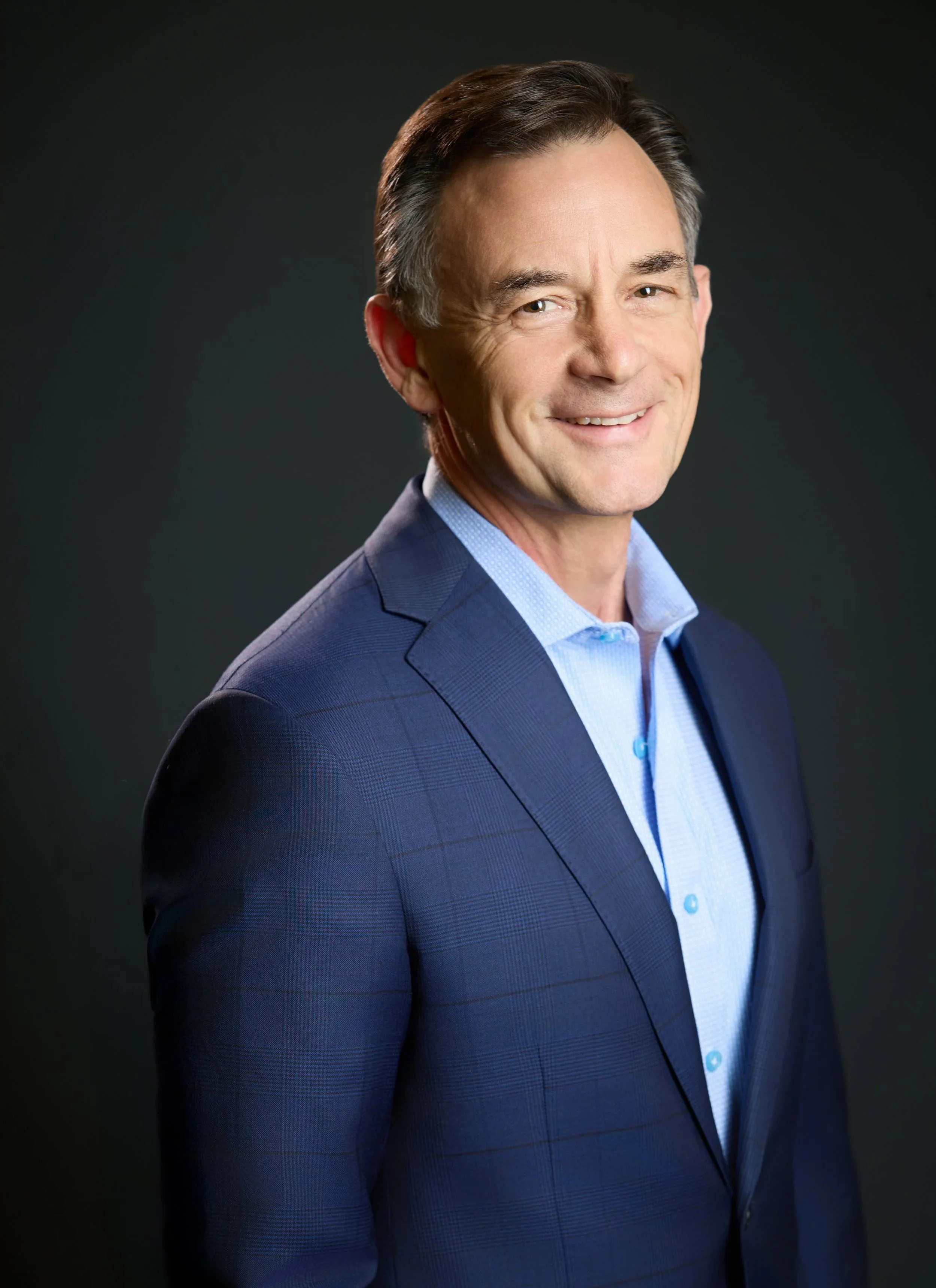 Dr. Russell Petrie wearing a blue suit and a light blue shirt, smiling against a dark background.