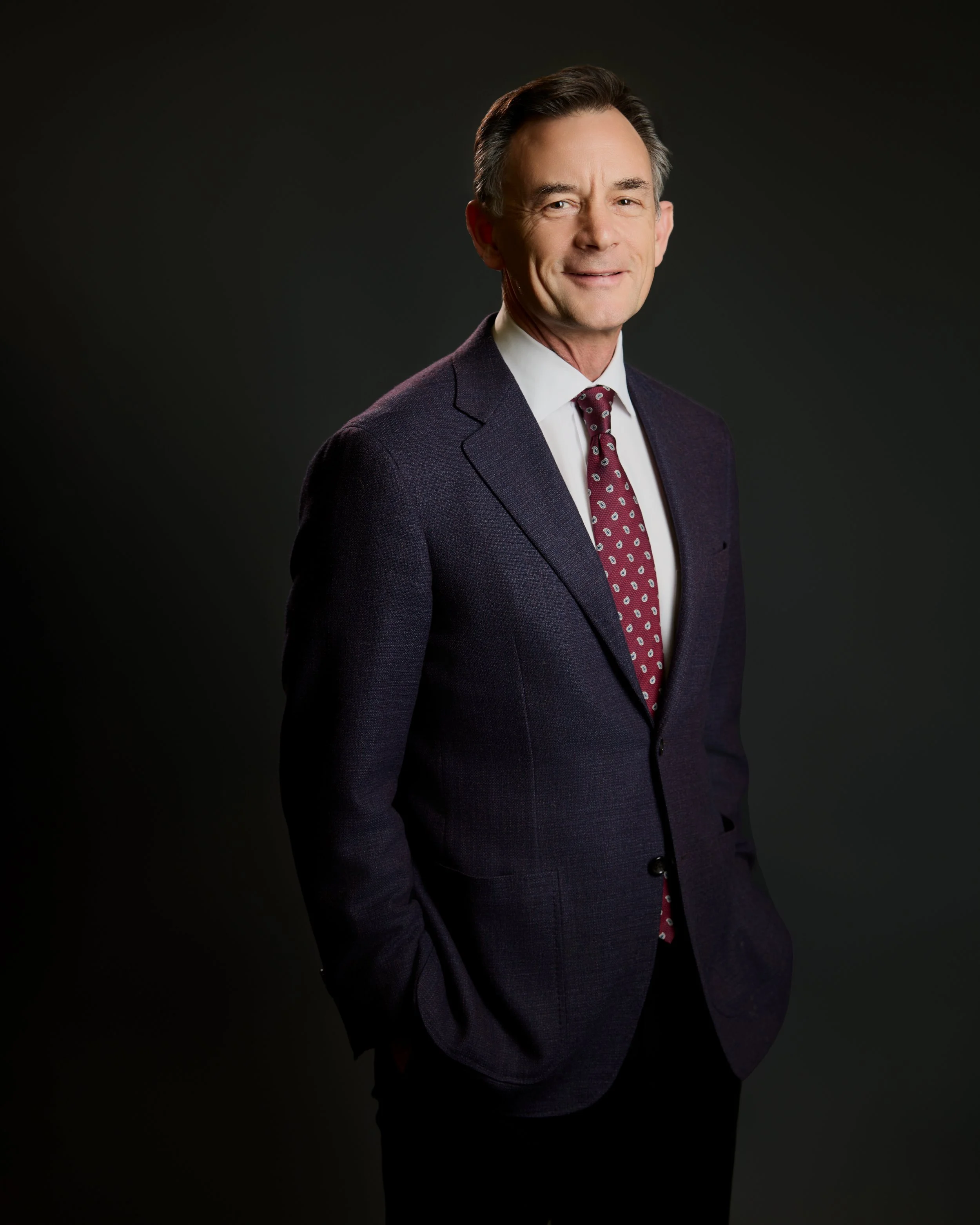 Dr. Russell Petrie wearing a dark suit, white shirt, and patterned maroon tie, standing against a black background.