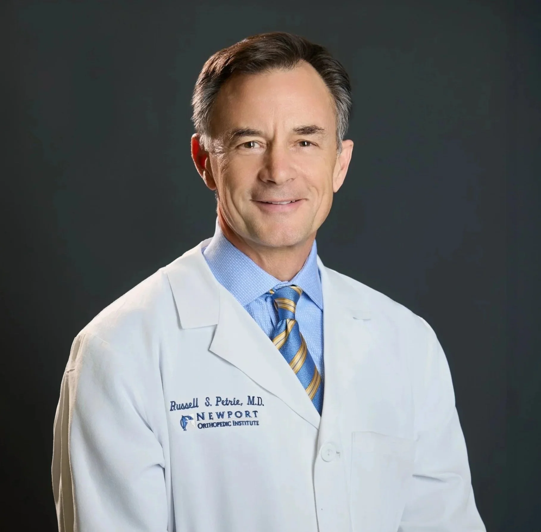 Headshot of Dr. Russell Petrie wearing a white lab coat with embroidered name and institution, blue shirt, and patterned tie, against a dark gray background.