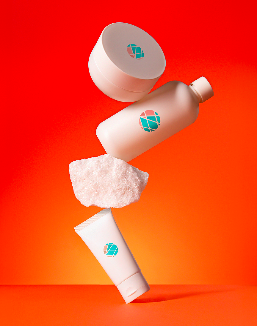 Four skincare or cosmetic products with Okeanos logo, two bottles, one jar, and one tube, arranged with a white pumice stone on an orange background.