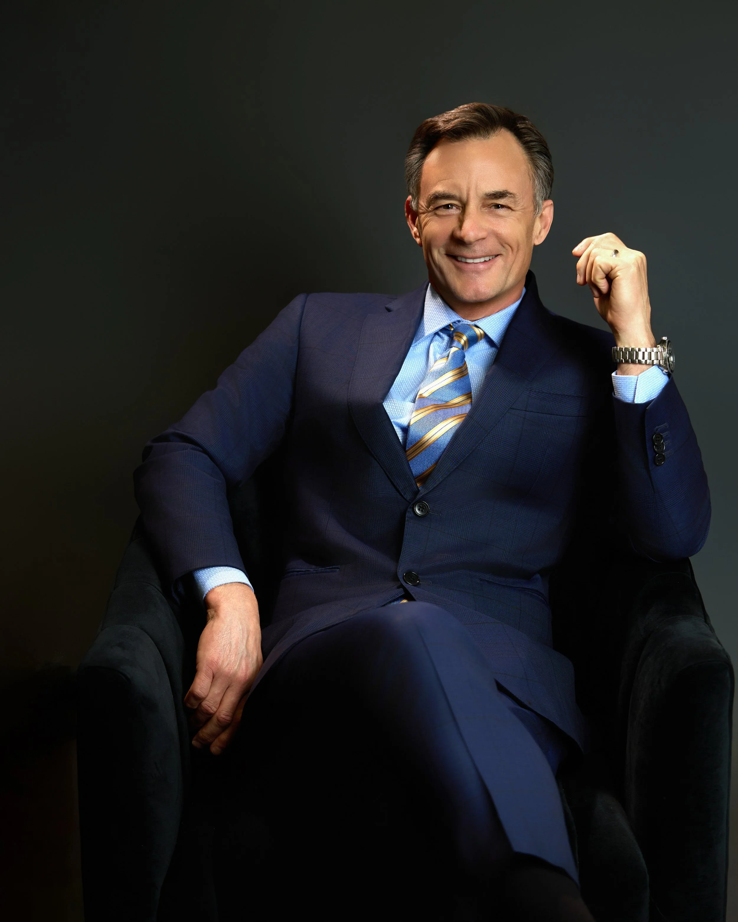 Dr. Russell Petrie wearing a blue suit with a light blue shirt and yellow striped tie, sitting confidently with one hand making a fist and the other resting on his leg, smiling in front of a dark background.