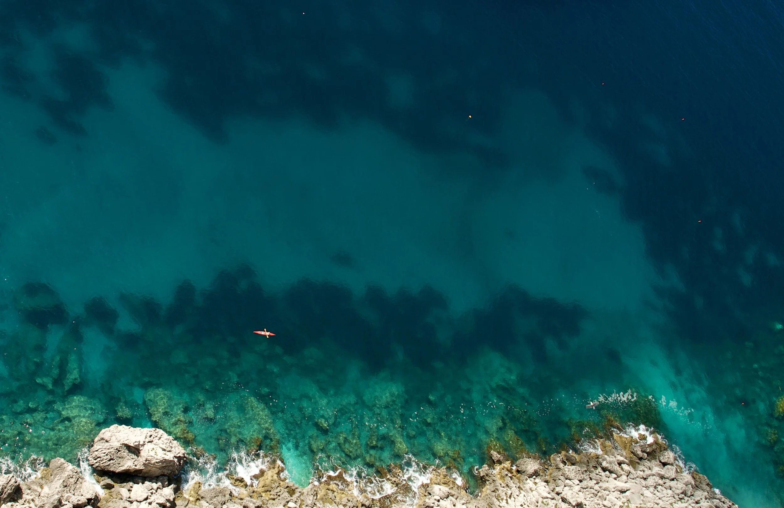 Aerial view of a rocky coastline with turquoise ocean waters and a small red boat island with two people on it.