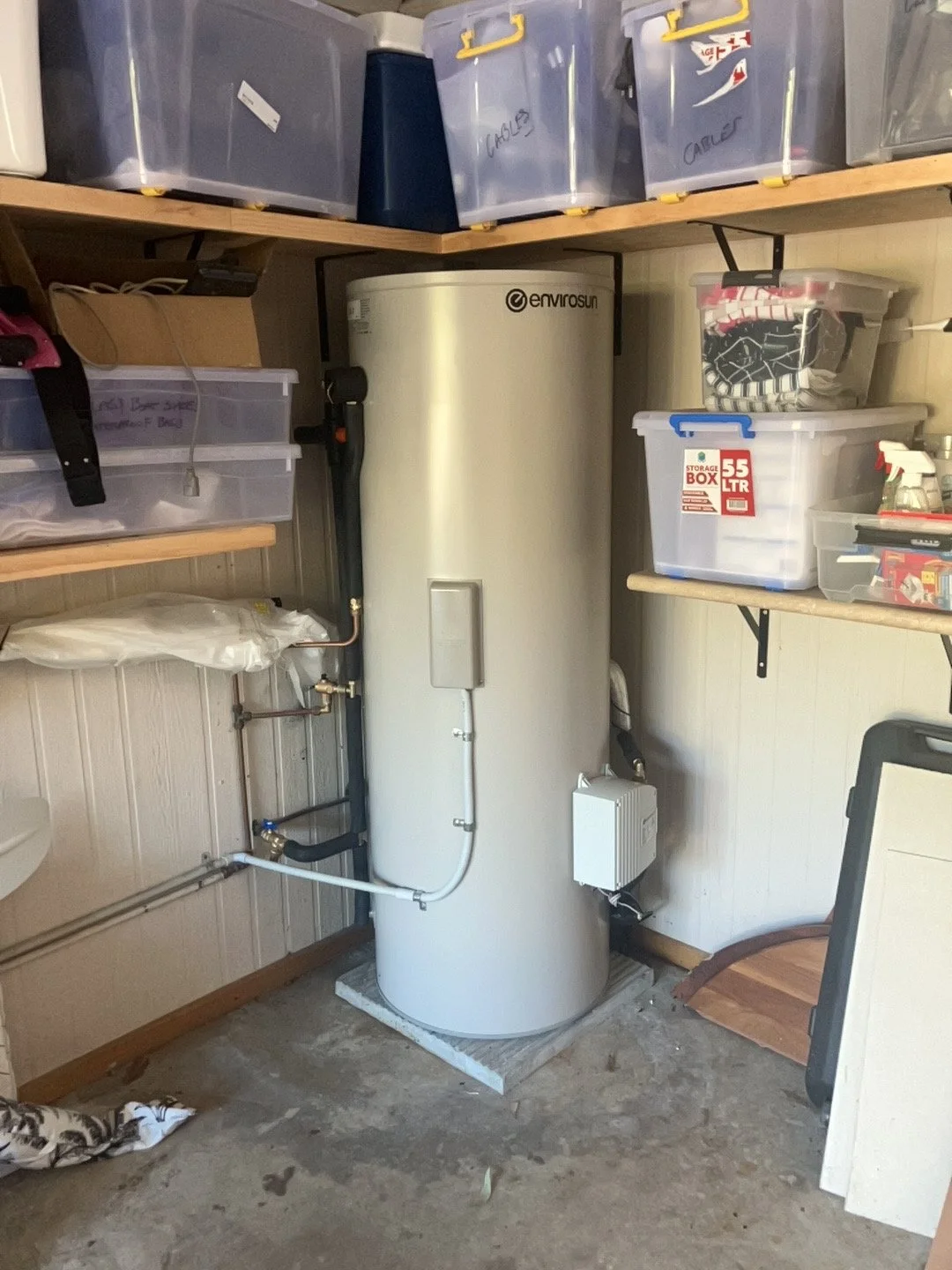 A tall, cylindrical water heater with a brand label 'envirosun' in a garage or storage room, surrounded by plastic storage bins on shelves.