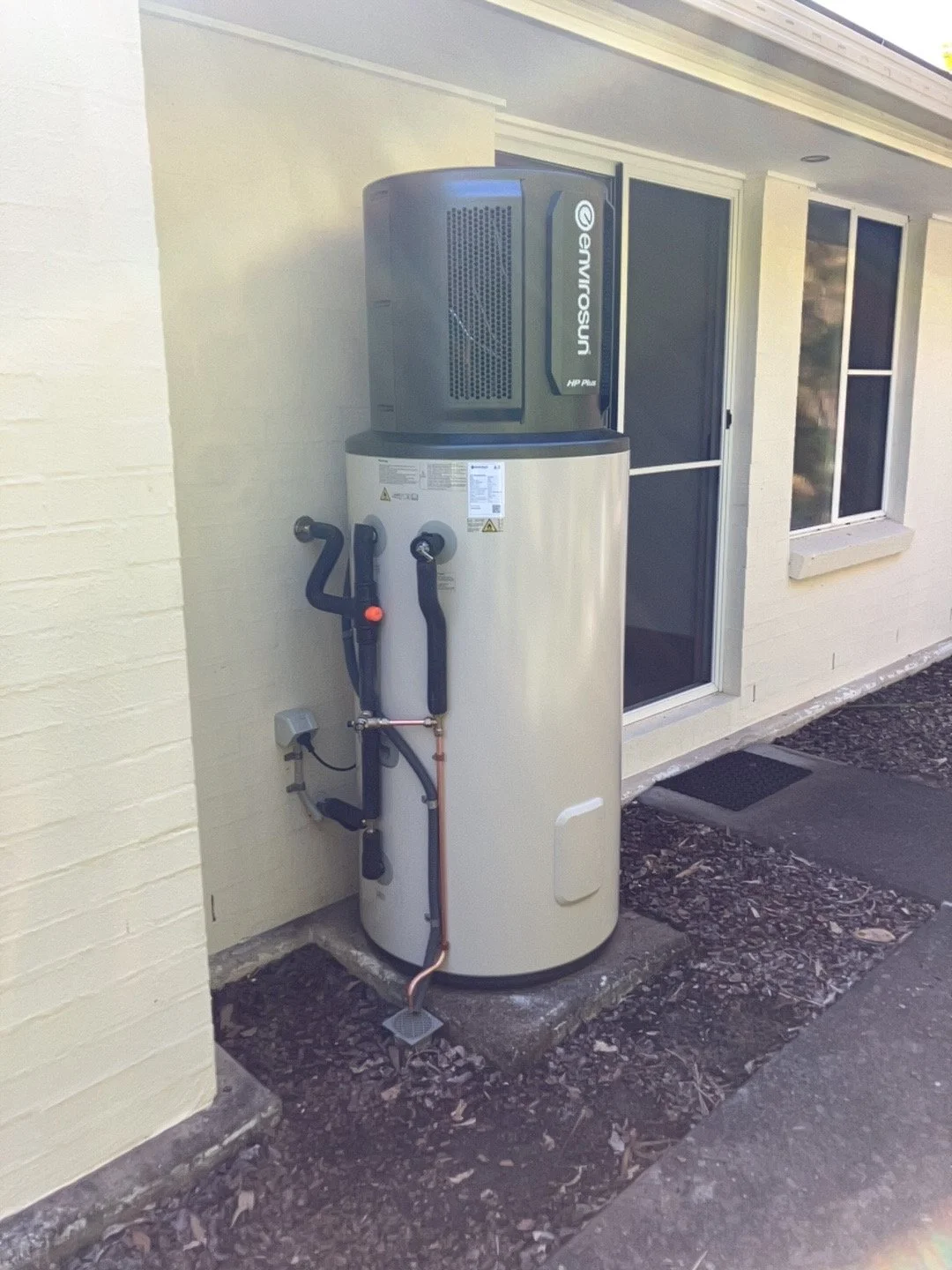 An outdoor heat pump system installed next to a house wall, featuring a tall white water heater tank and a gray air source heat pump on top.