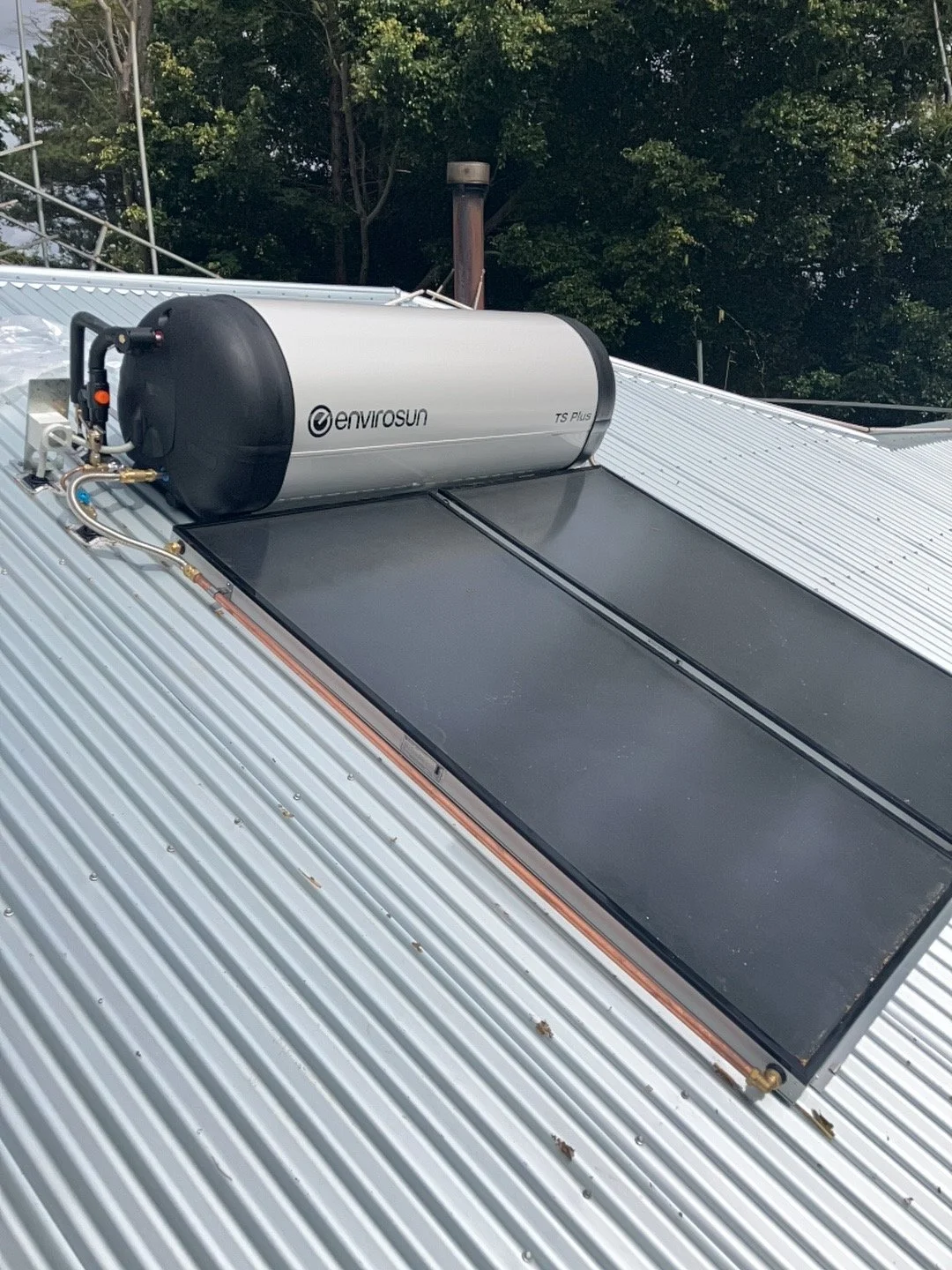 A solar water heater installed on a metal roof, with an EnviroSun solar collector, against a background of green trees.