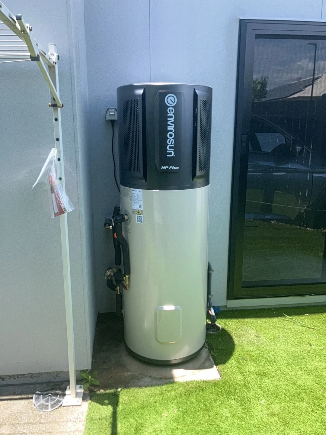 Outdoor air-to-water heat pump system in a backyard setup, with a black top section and a light-colored cylindrical tank below, situated next to a gray wall and a black glass door, on a patch of green grass.
