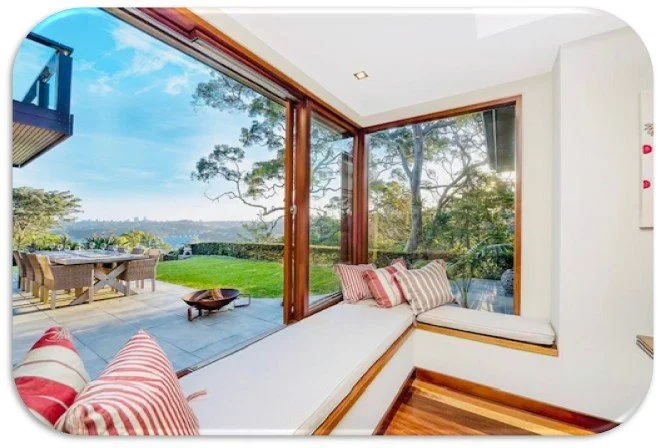 Interior view of a sunroom with large glass windows and a cozy window seat with striped cushions, overlooking a lush green outdoor patio and trees.