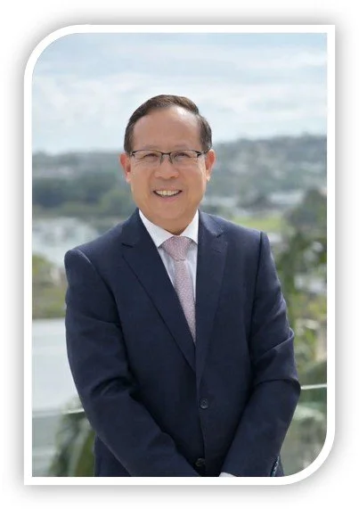 Man in a navy suit, white shirt, and light-colored tie standing outdoors with a cityscape and cloudy sky in the background.